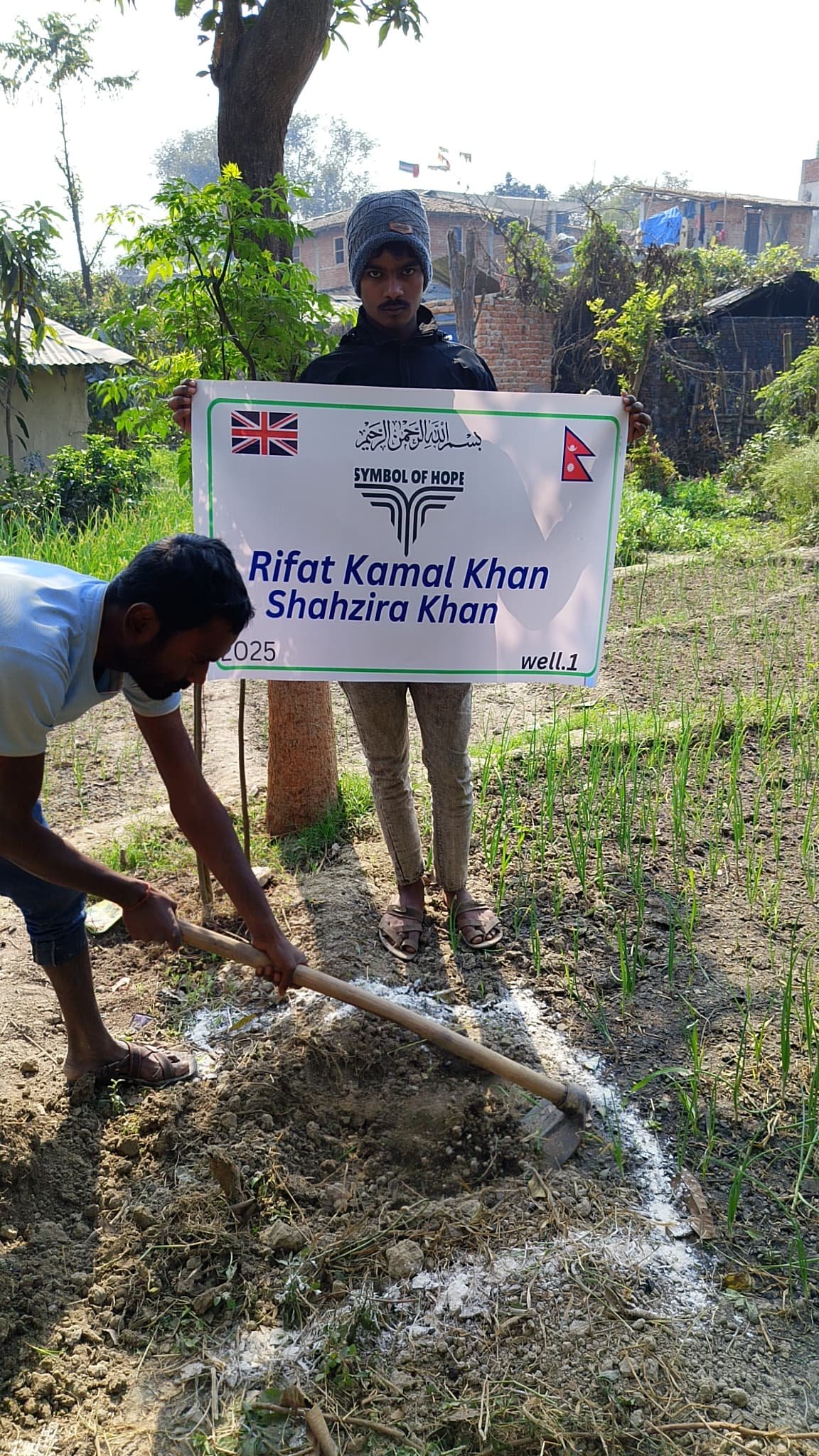 Two men in a field, one holding a sign that reads 'Symbol of Hope' and names, the other digging with a tool, in front of a rural backdrop with plants.