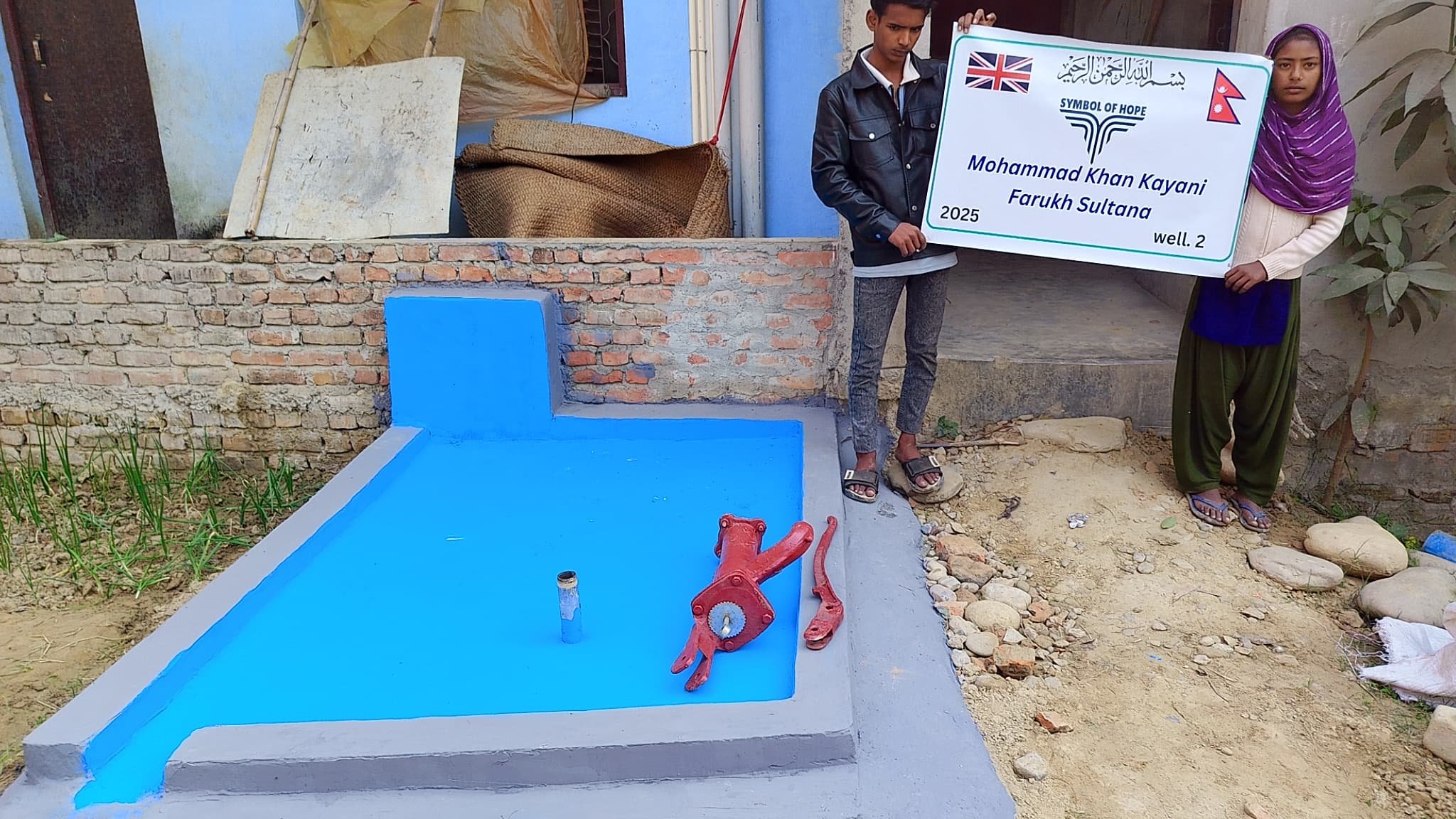 Two people holding a sign near a newly constructed water pump on a blue platform, indicating a charity project or donation initiative.
