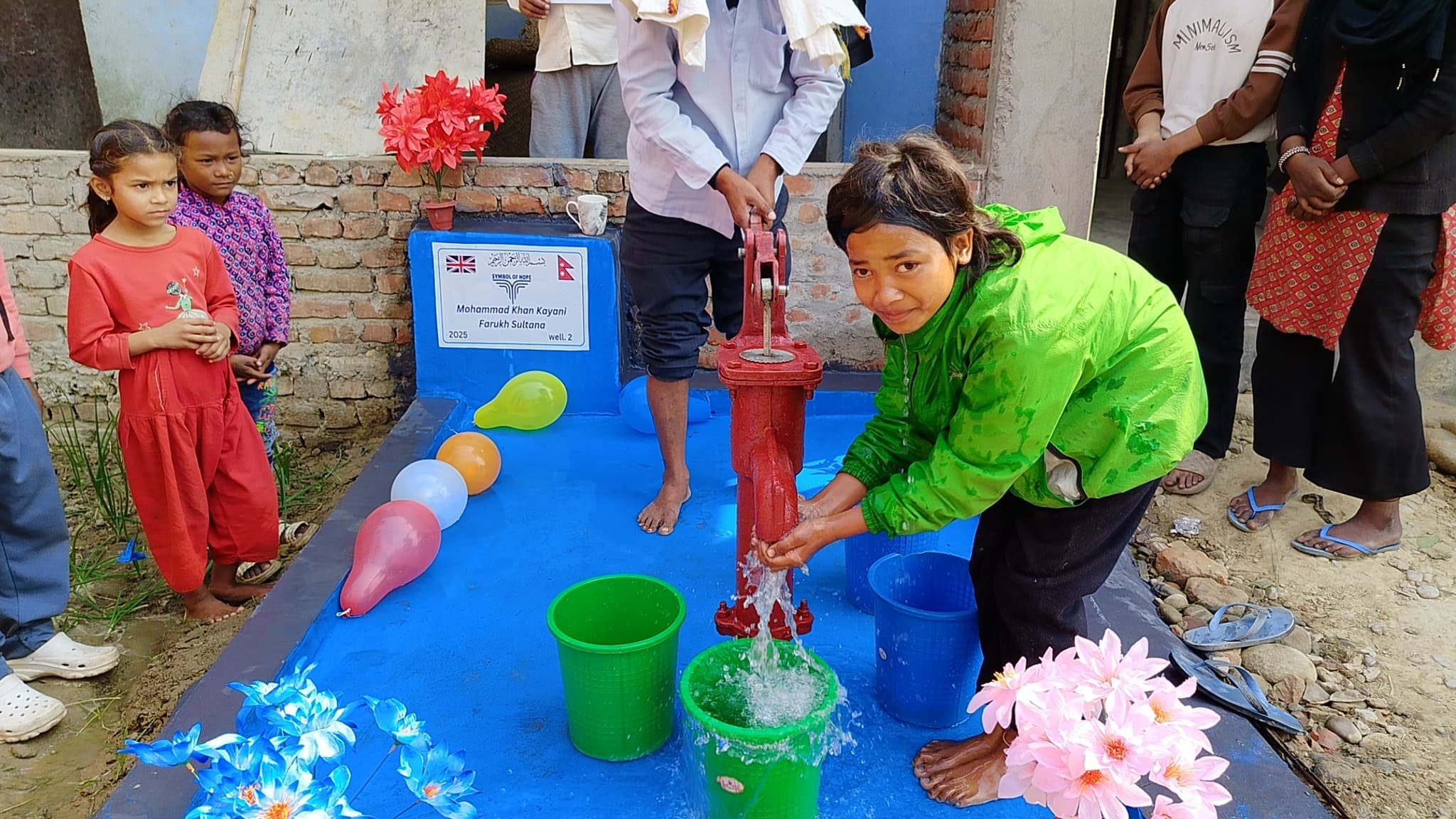 Children using a hand pump to fill buckets with water in a rural setting with colorful balloons and flowers.