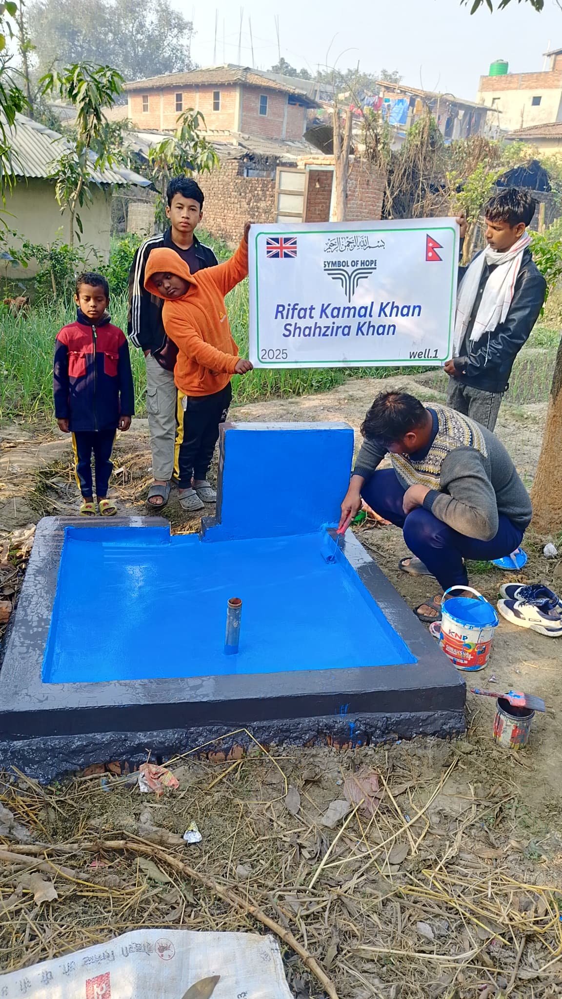 Group of people around a newly constructed blue water well in a rural area, with a sign displaying U.K. and Nepal flags and names Rifat Kamal Khan and Shahzira Khan.