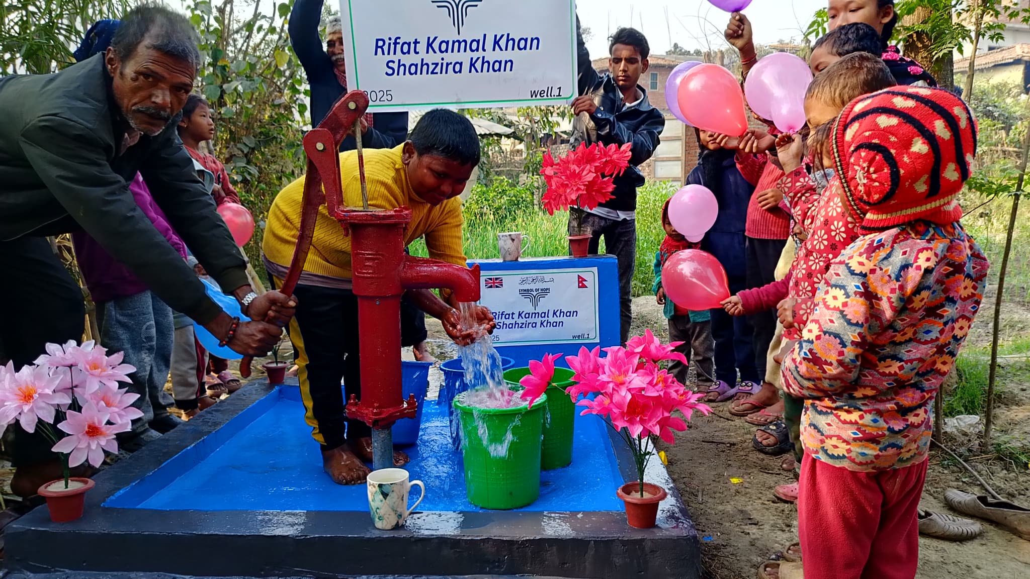 Group of people, including children with balloons, gathered around a water pump with flowers and signage.