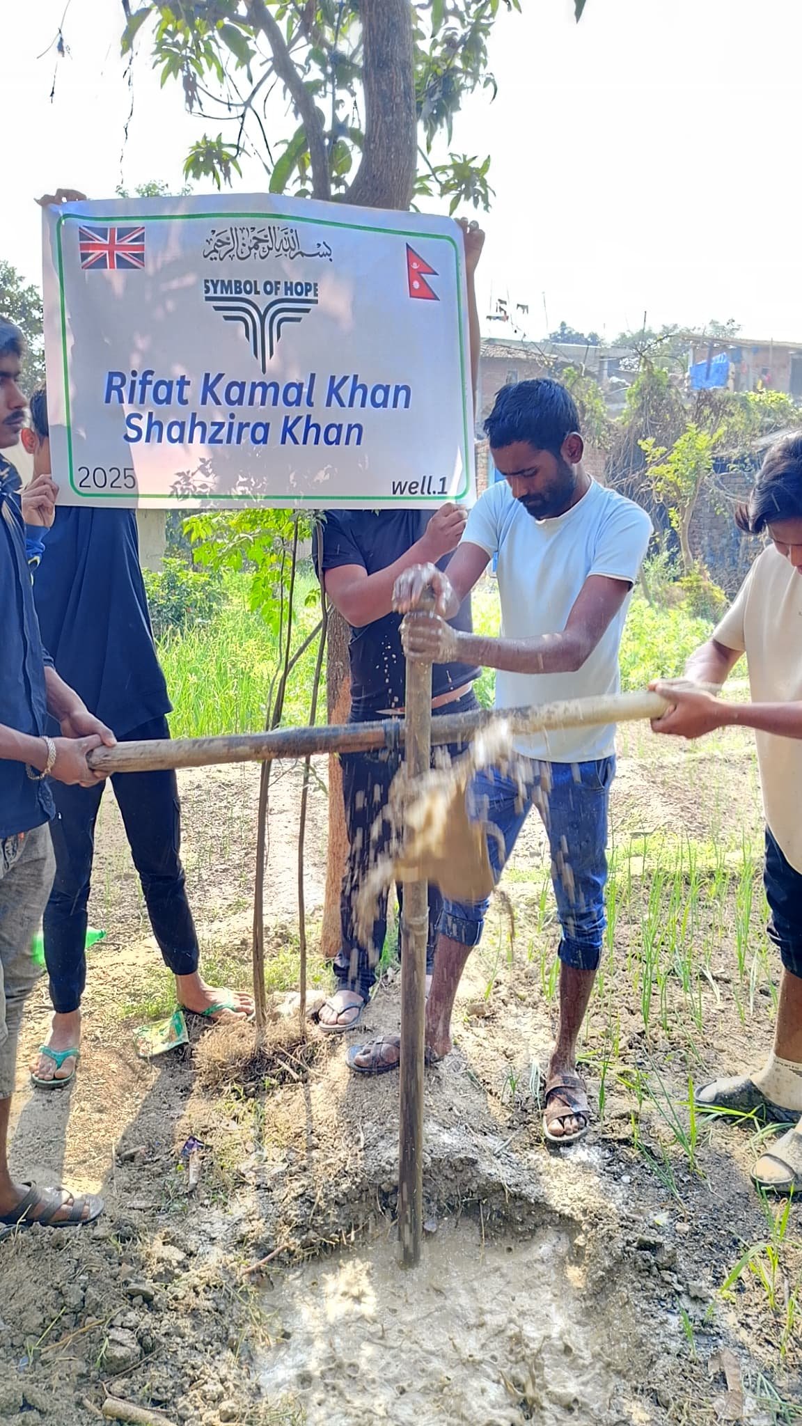 People using a manual pump to drill a well, with a banner overhead containing text and flags, surrounded by greenery.