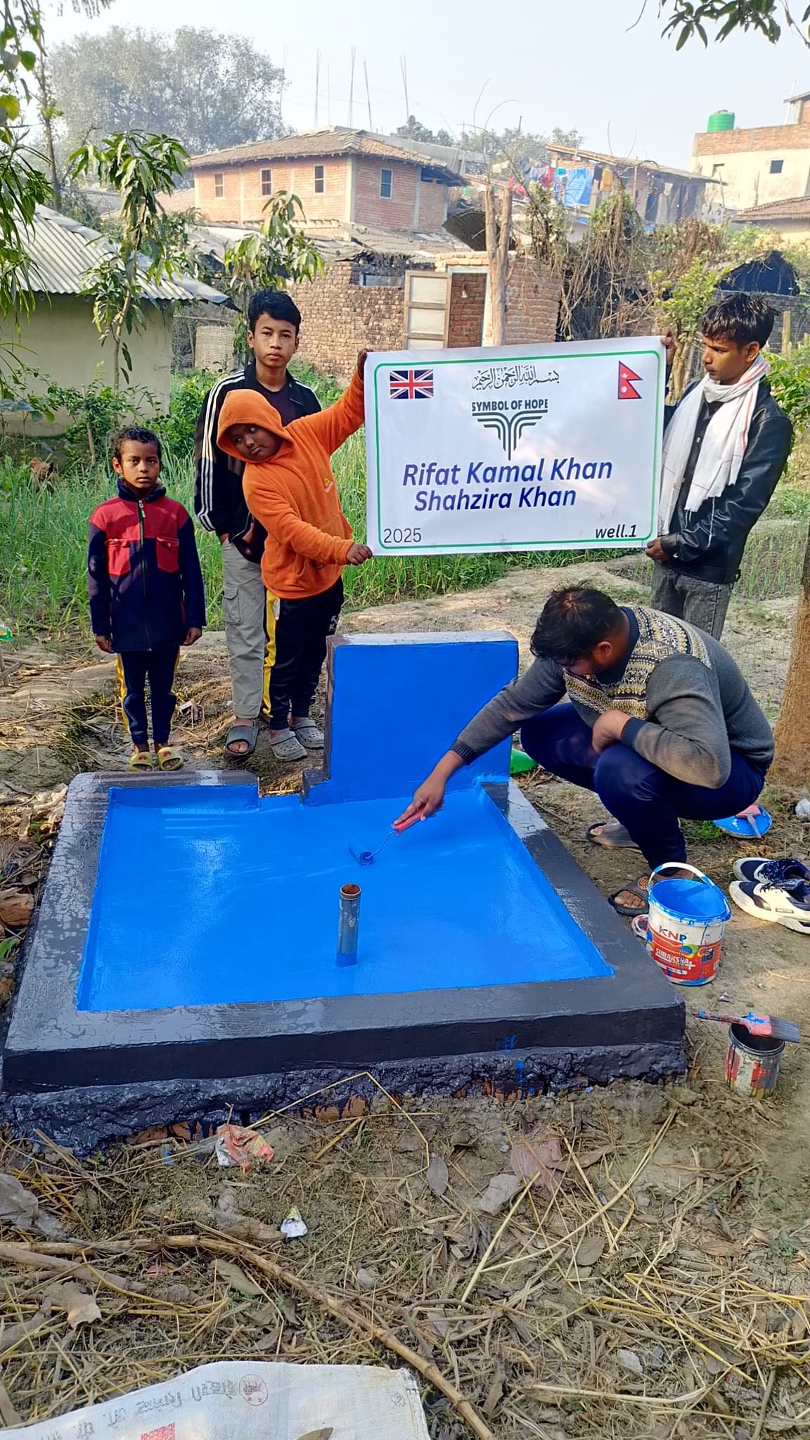 Group of people near a newly painted blue well, holding a sign with names and flags.