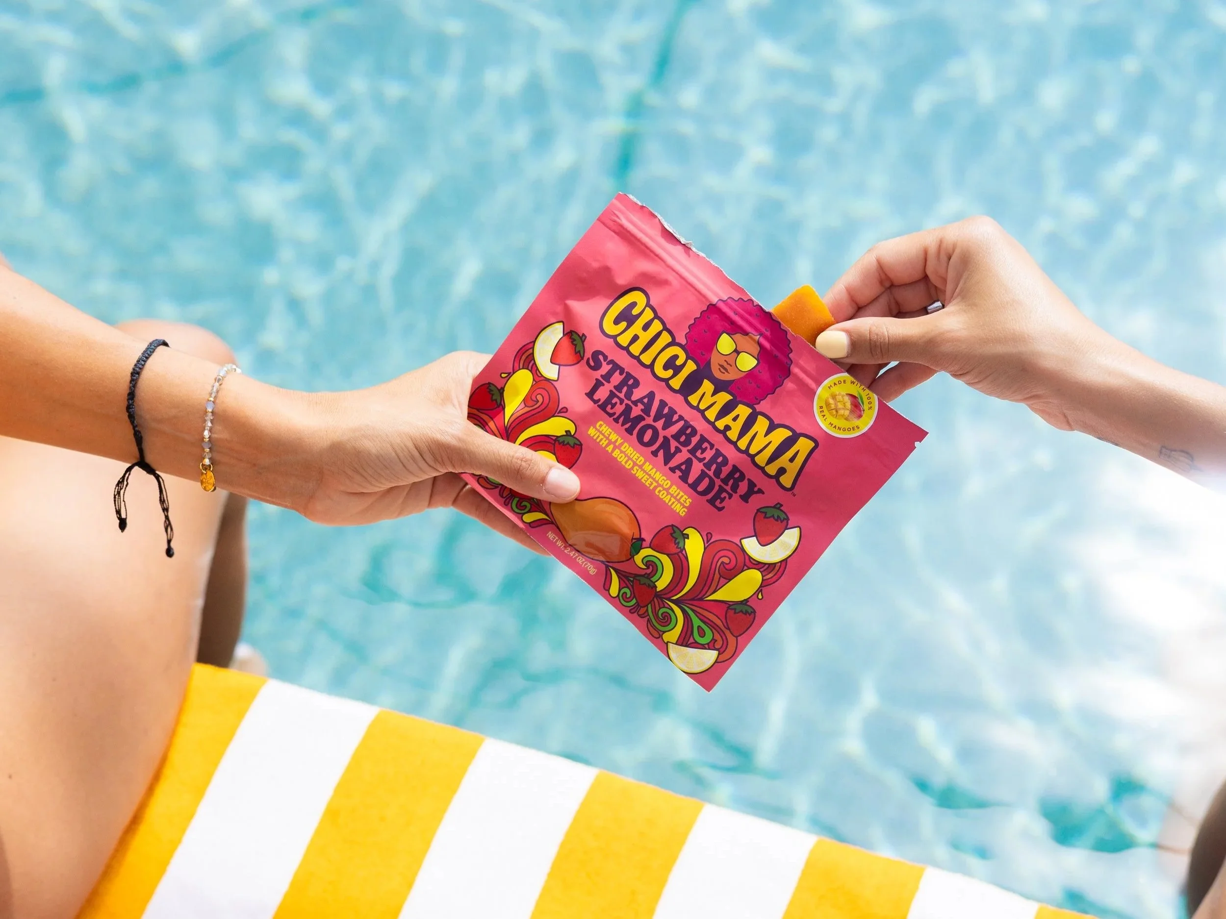 Hand offering Chici Mama snack bag over pool water on yellow striped towel, summer brand content by Gabe Gonzalez