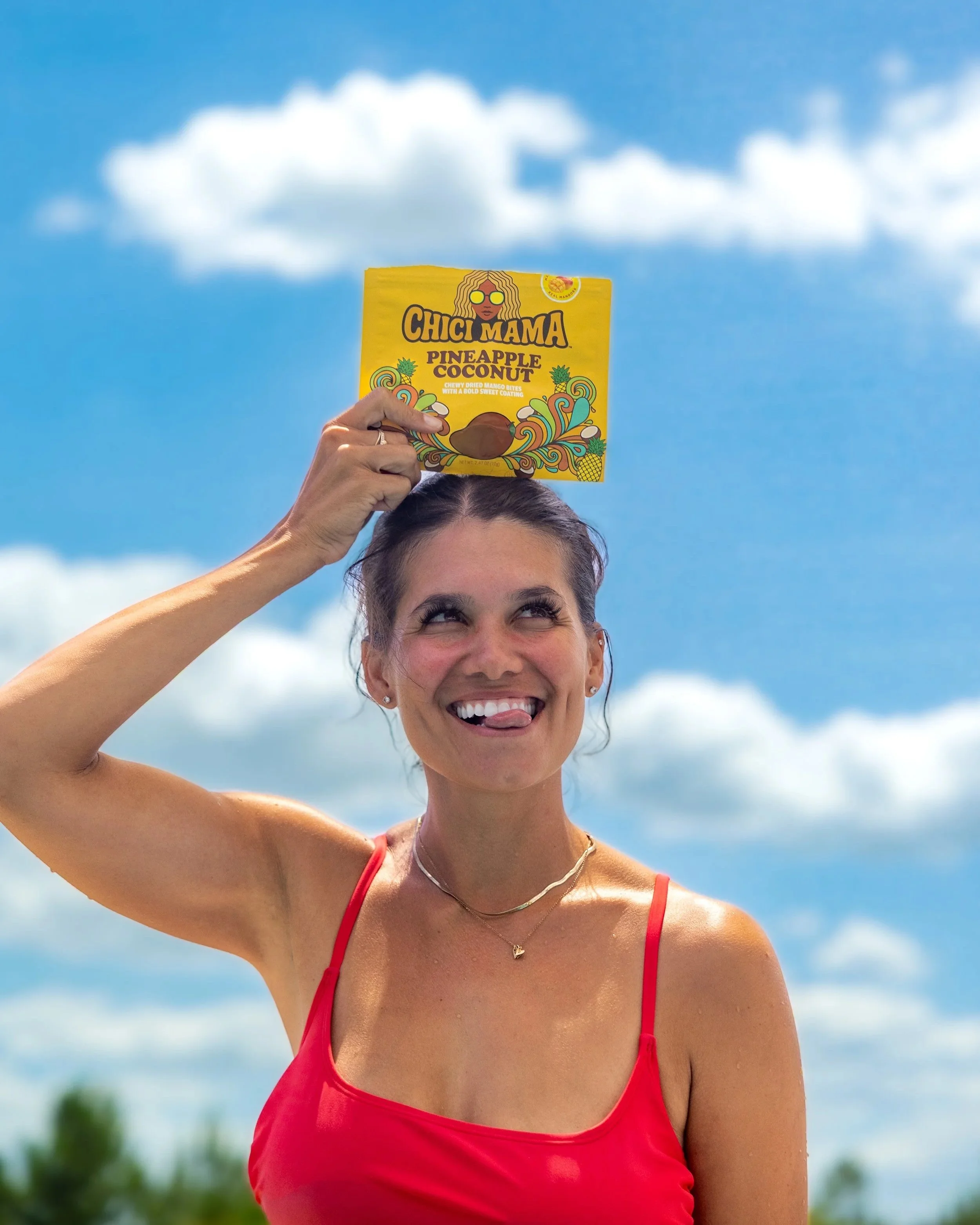 Woman in red swimsuit smiling holding Chici Mama snack bag against blue sky, joyful summer campaign by Gabe Gonzalez