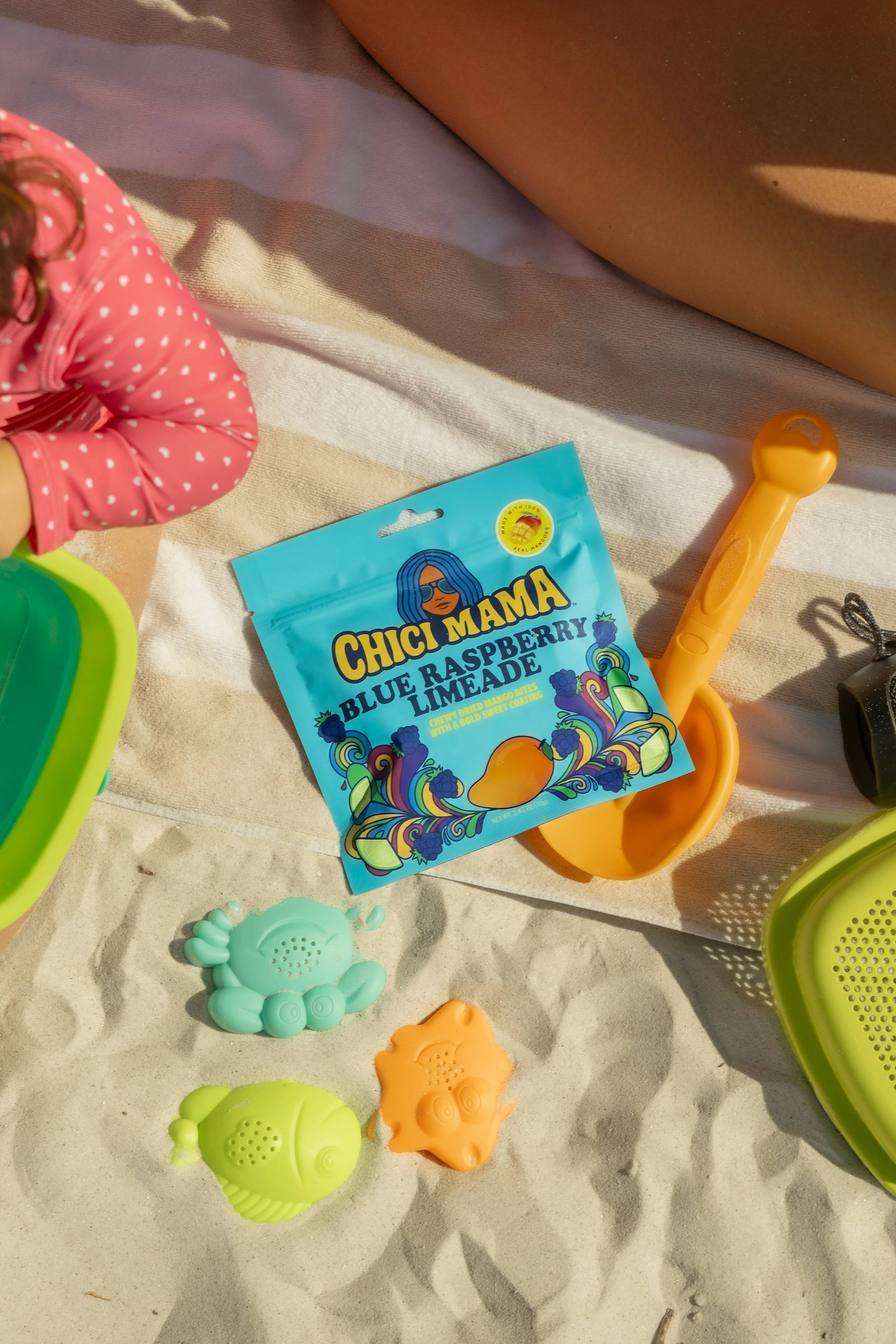 Toddler with Chici Mama snack bag surrounded by colorful sand toys at beach, family summer photography by Gabe Gonzalez