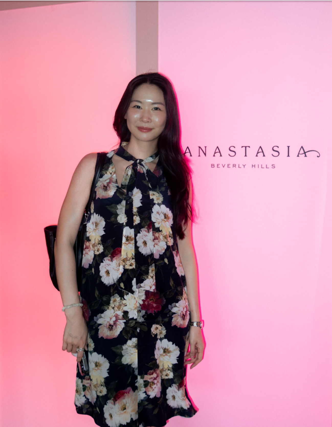 A woman with long dark hair wearing a floral dress and carrying a purse, standing in front of a pink-lit wall with the text 'Anastasia Beverly Hills'.