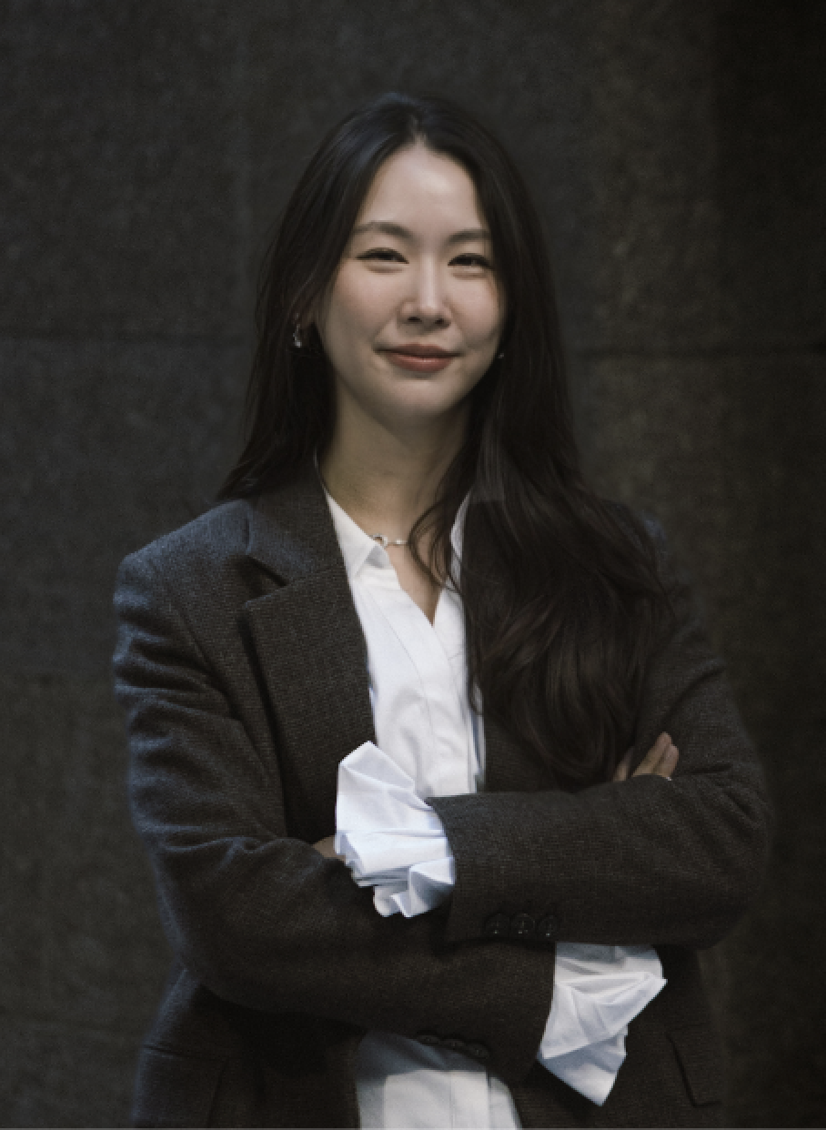 Portrait of a woman with long dark hair, wearing a dark blazer and white blouse, standing against a dark background with arms crossed and a subtle smile.