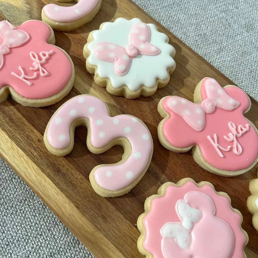 Decorated sugar cookies on a wooden platter, featuring pink, white, and polka-dotted icing with butterfly and heart designs, some cookies with the name 'Kya' written on them.