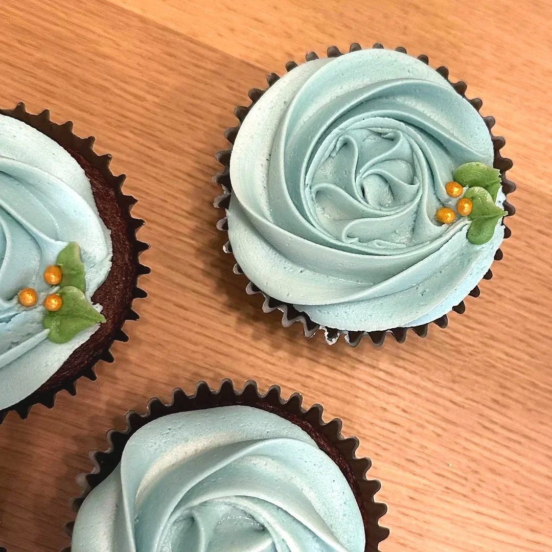 Three cupcakes with light blue frosting and gold and green decorative accents on top, positioned on a wooden surface.