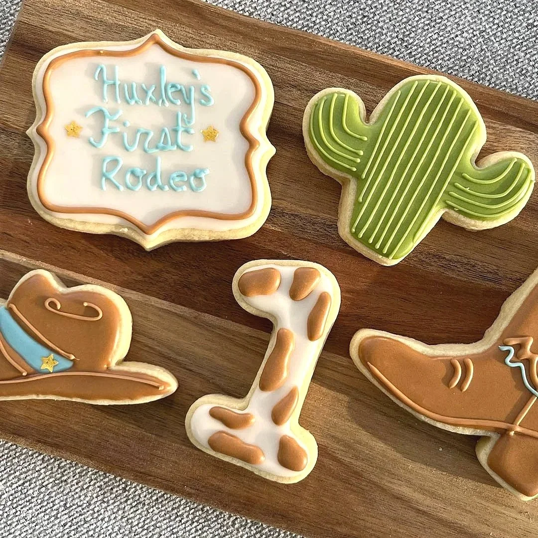 Cookies decorated with Western and cowboy themes on a wooden board, including a sheriff's badge with the message "Huxley's First Rodeo," a cowboy hat, a cowgirl boot, a cactus, and a horse shoe.
