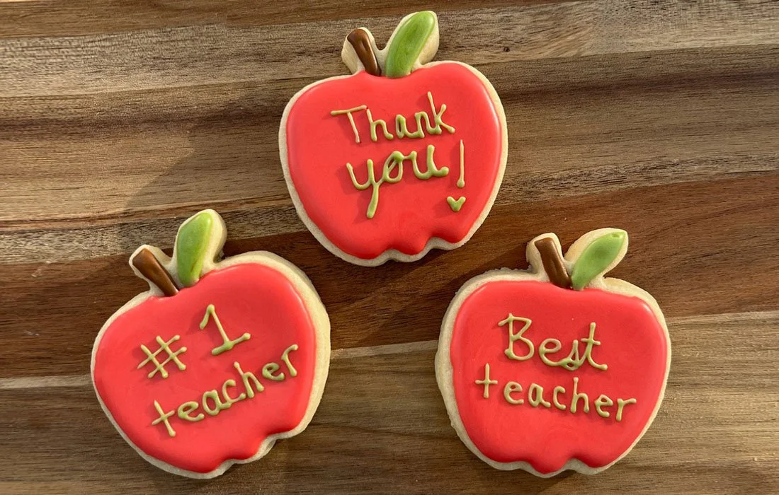 Three decorated cookies in the shape of red apples with green leaves and brown stems, with messages written in yellow icing: "Thank you!", "#1 teacher", and "Best teacher" on a wooden surface.