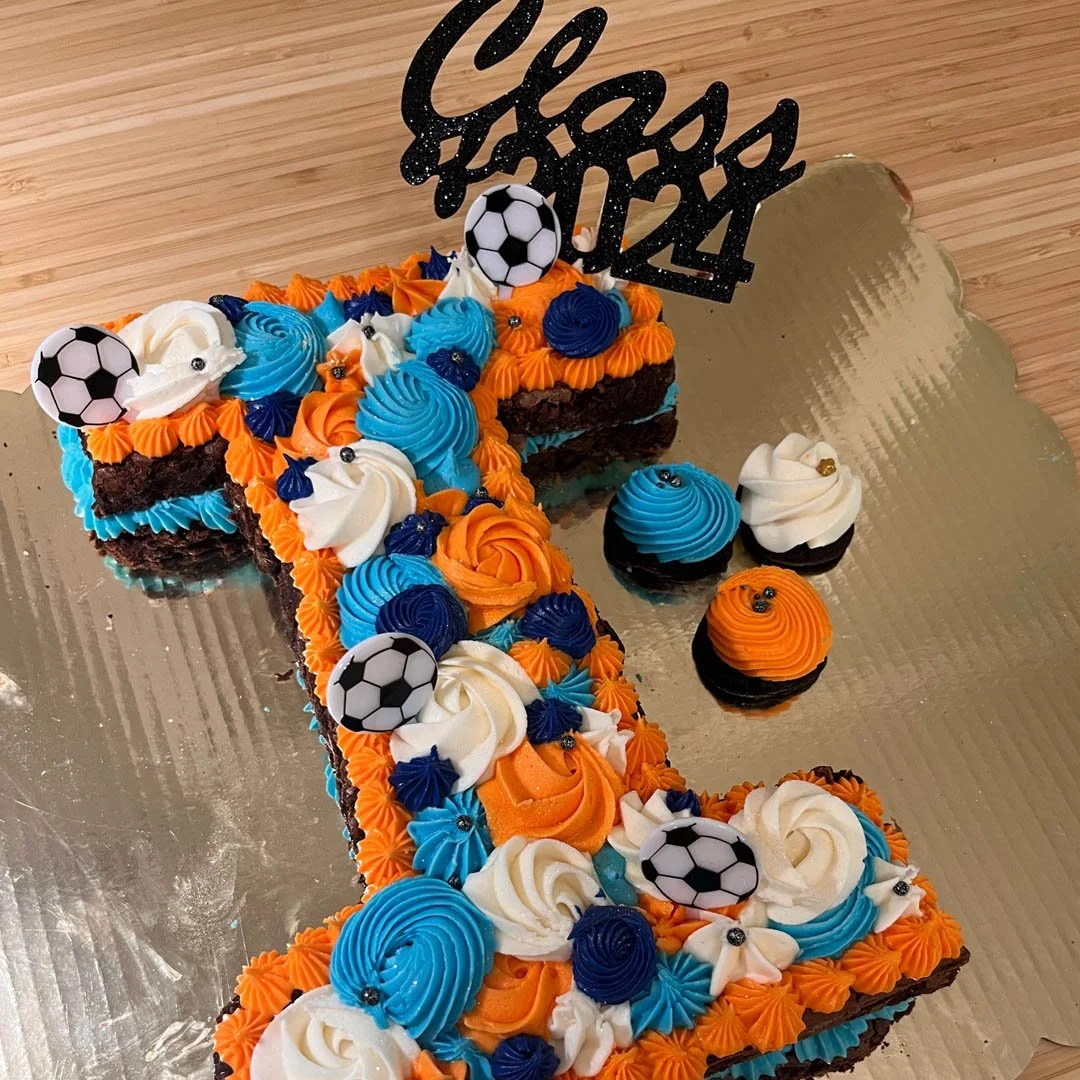 Decorative birthday cake shaped like the number 1, with orange, blue, and white frosting, soccer ball toppers, and a "Happy Birthday" topper.