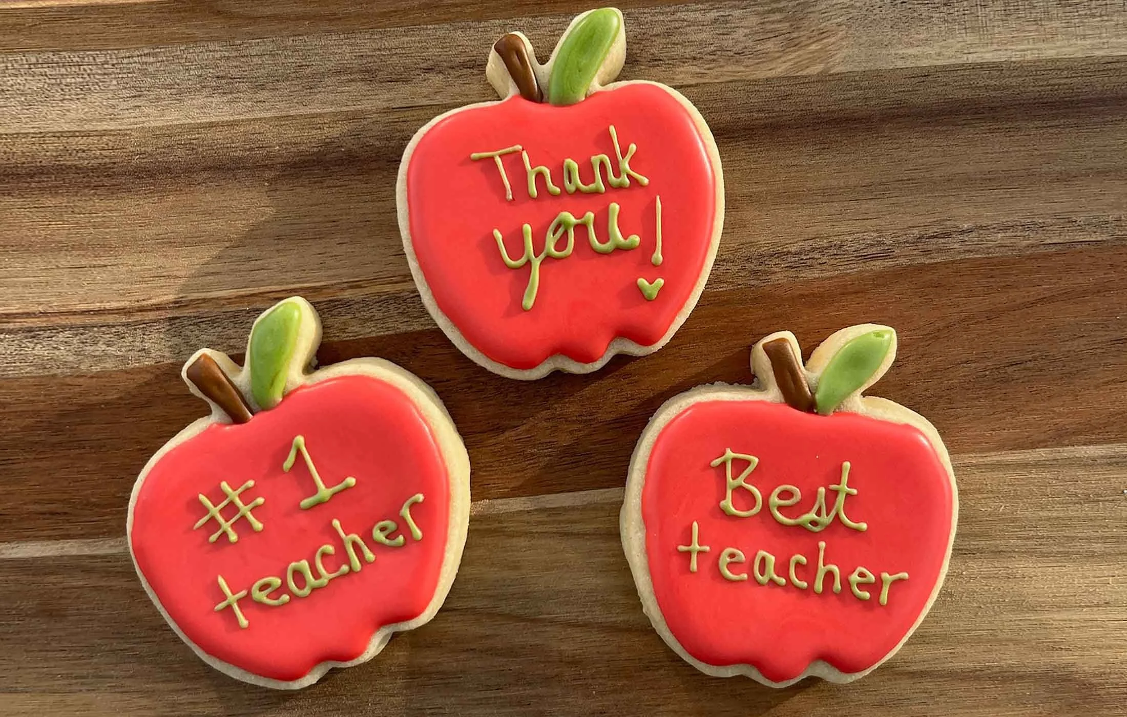Apple-shaped cookies with red icing, decorated with green and brown icing leaves and words like "Thank you!" and "Best teacher" written in yellow icing, placed on a wooden surface.