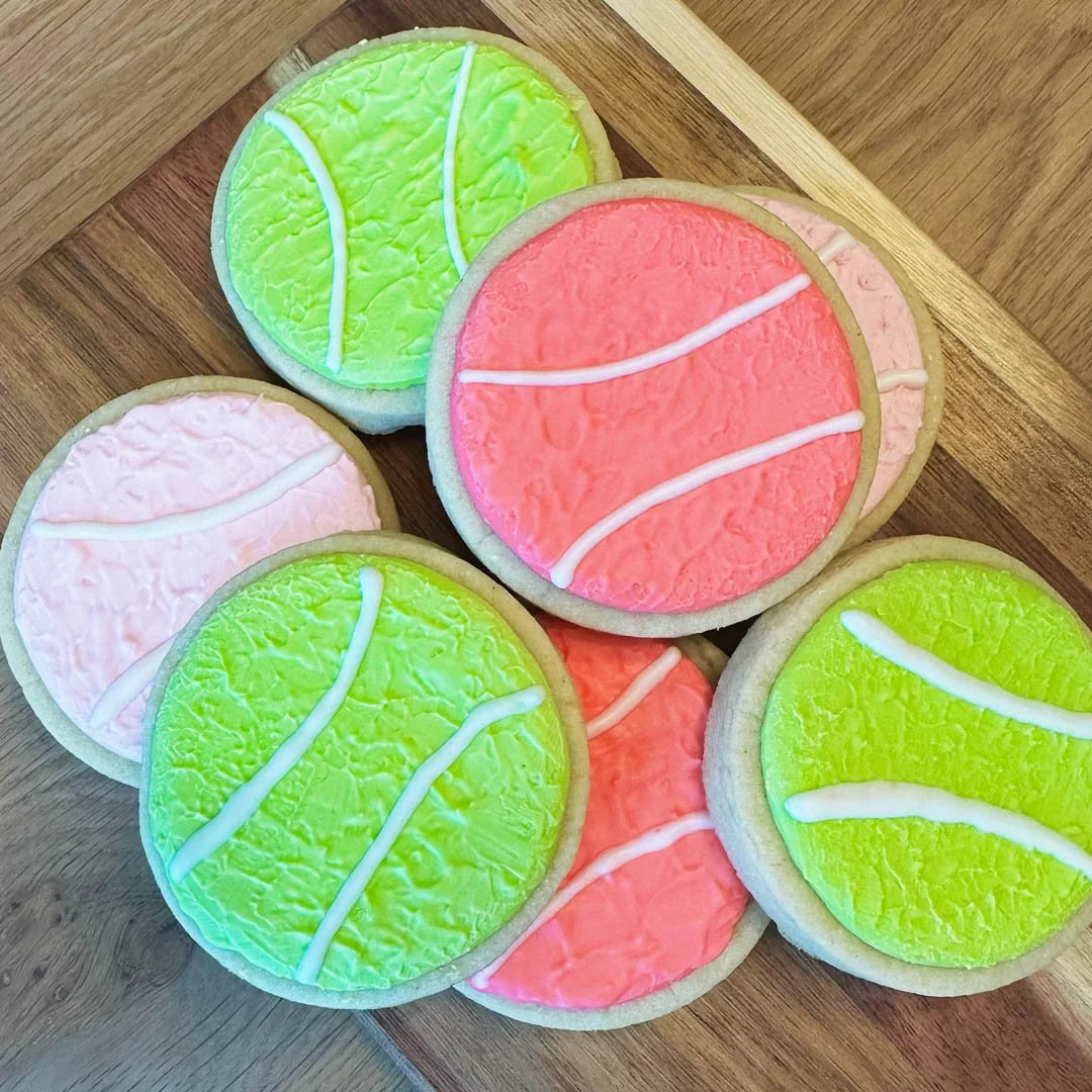 A collection of tennis ball-shaped sugar cookies with green, pink, and white icing, decorated to resemble tennis balls, placed on a wooden surface.