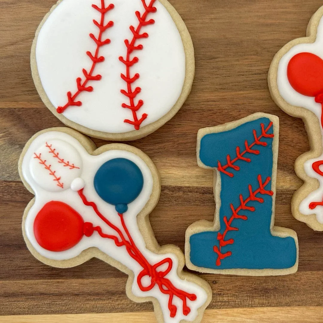 Decorated baseball-themed cookies on a wooden surface, including a white baseball with red stitching, a blue and red balloon with baseball stitching, and a blue number 1 with red stitching.
