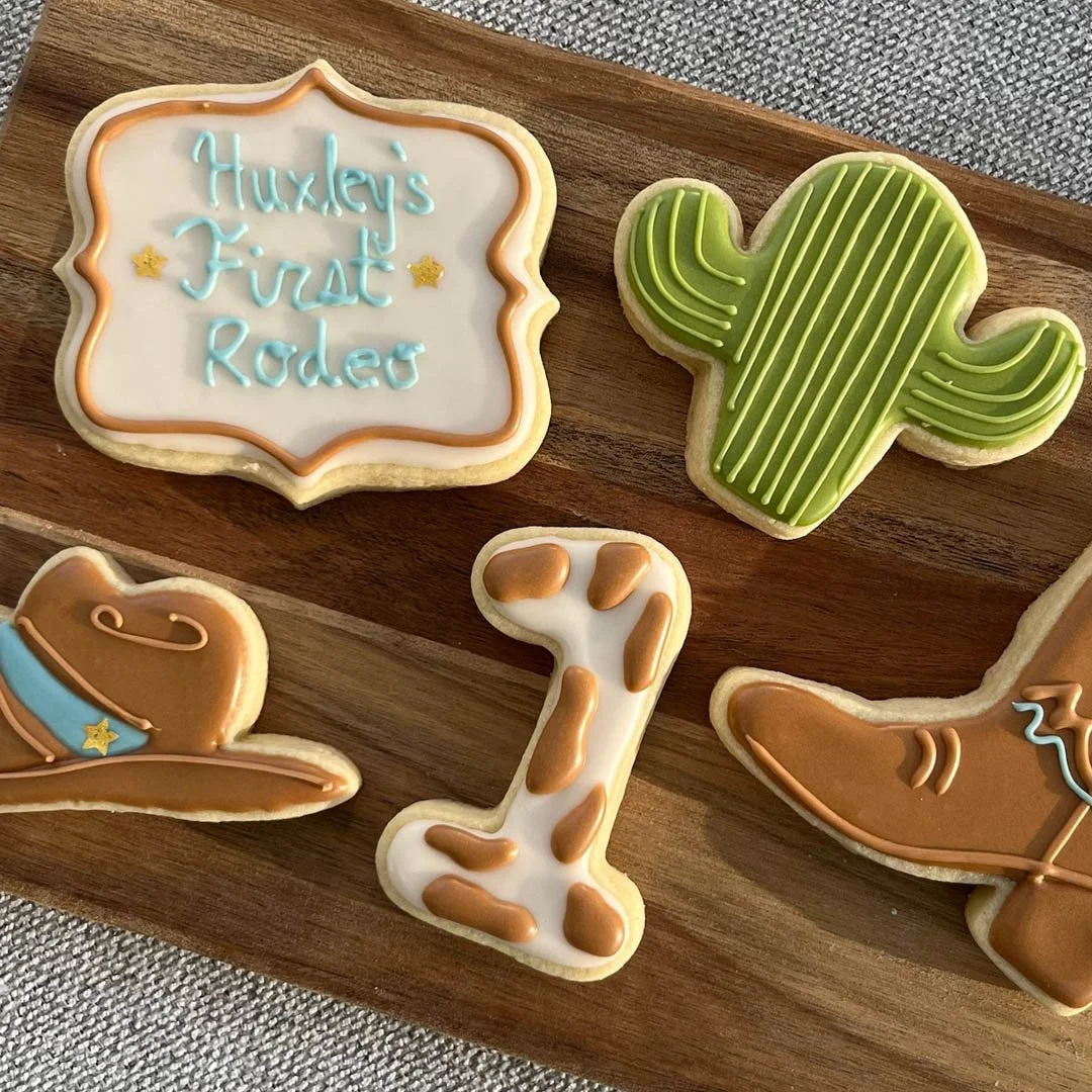 Decorated sugar cookies themed around Wild West and cowgirl elements. One cookie has a sign with the text 'Huxley's First Rodeo' in light blue icing, another is shaped like a cactus with green icing and lines, a third shaped like a cowboy hat with li
