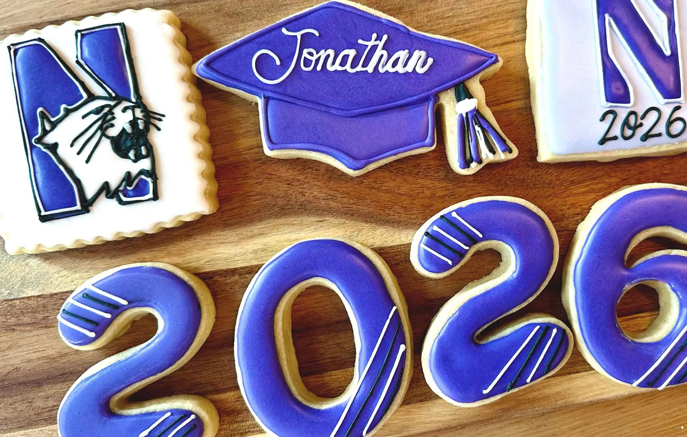 Decorated cookies with purple and white icing, featuring a graduation cap with the name 'Jonathan' and the year '2026' written on them, placed on a wooden surface.