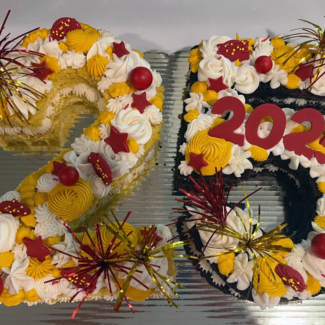 Decorative cake shaped like the number 2023 with yellow and white frosting, red star and ball decorations, and red number candles, celebrating the New Year.