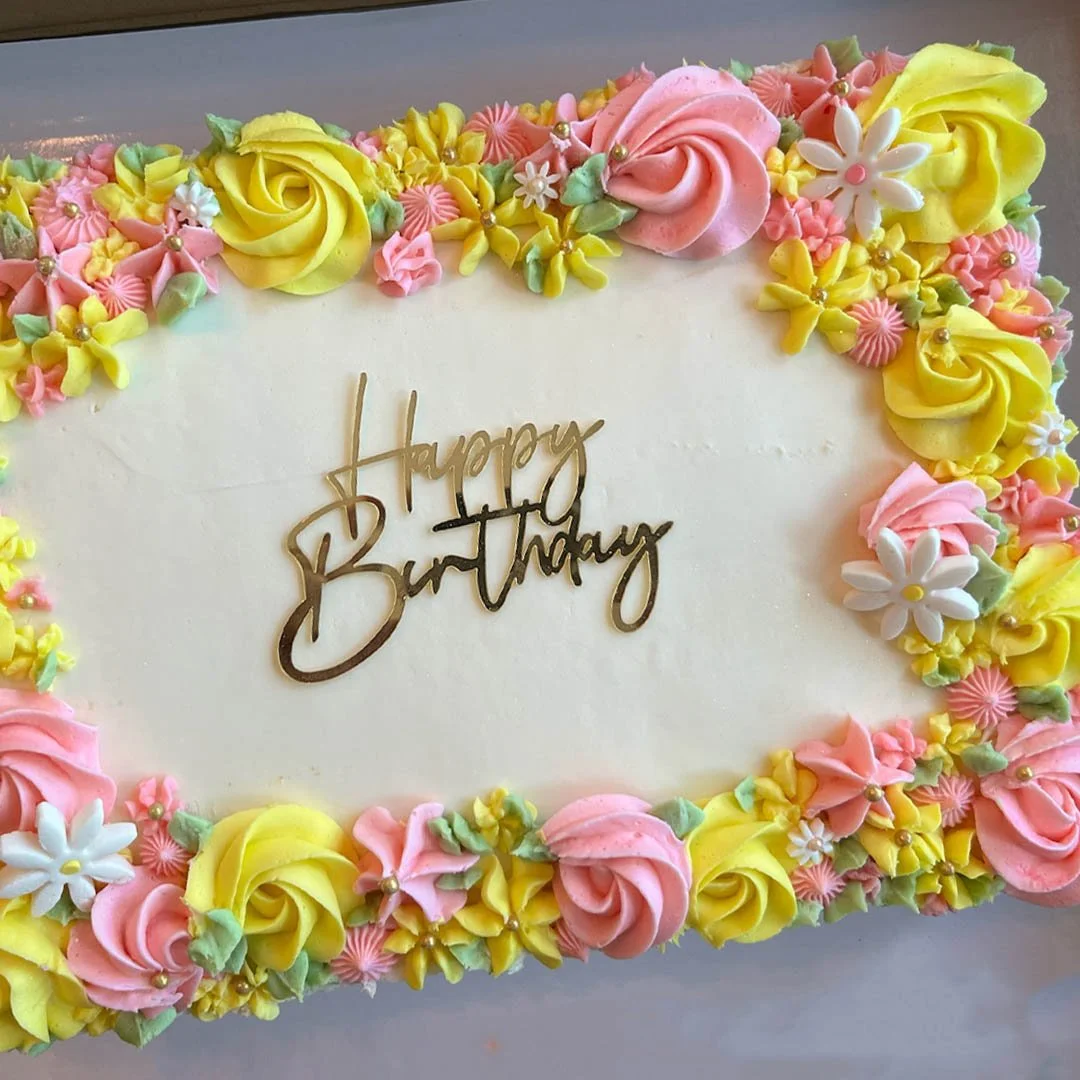 A decorated birthday cake with pink, yellow, and white fondant flowers around the edges and a gold 'Happy Birthday' topper in the center.