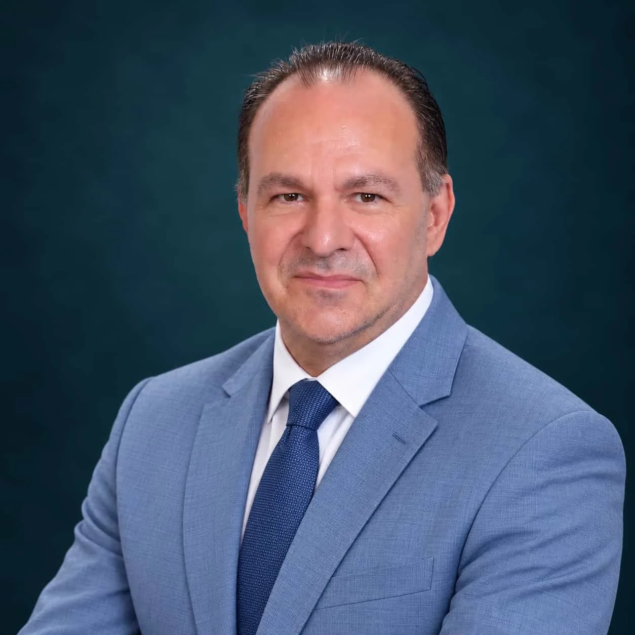 Professional headshot of a man in a light blue suit and navy tie against a dark blue background