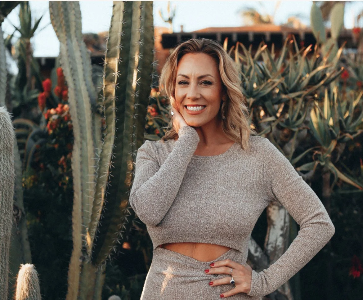 A woman with blonde hair smiling outdoors among cacti and succulents wearing a beige long-sleeve dress with a cut-out at the waist.