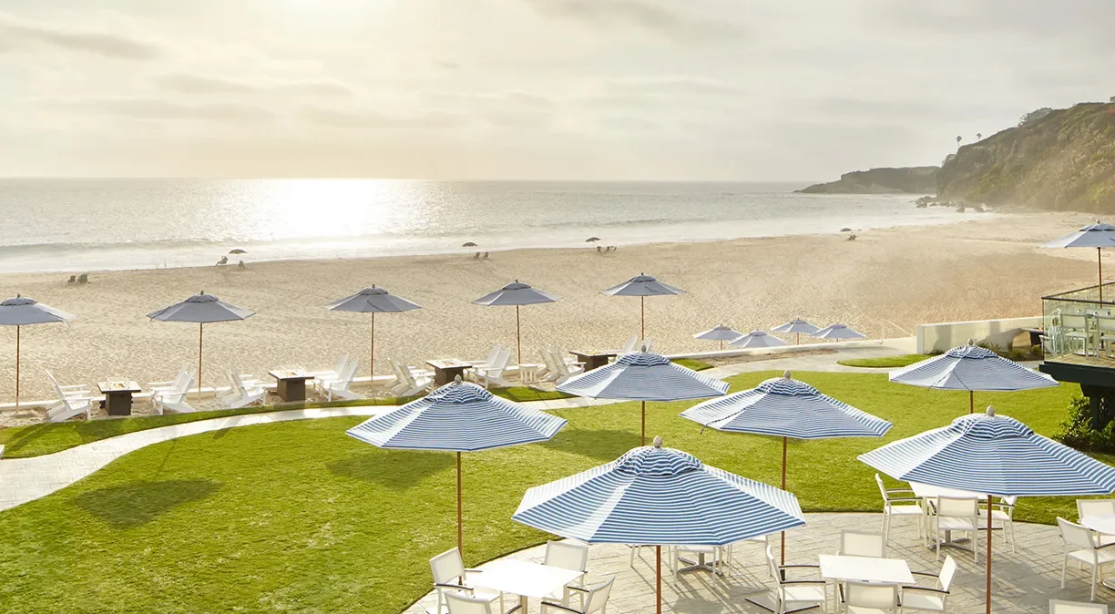 Beachside outdoor seating area with umbrellas, tables, and chairs overlooking sandy beach and ocean at sunset.