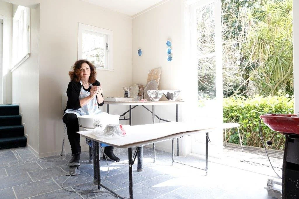 A woman in a black and white apron stands in a bright, airy kitchen with large windows overlooking greenery. She holds a dark object in her hands, possibly a piece of pottery, and there are various pottery items and tools on the tables around her.