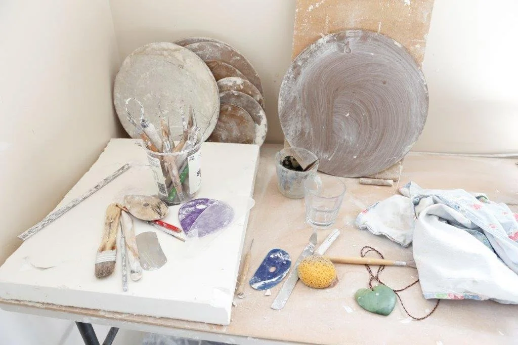 A cluttered artist's workspace with pottery wheels, stoneware pieces, paintbrushes in a jar, various tiles, a sponge, a glass of water, and a cloth on a table.