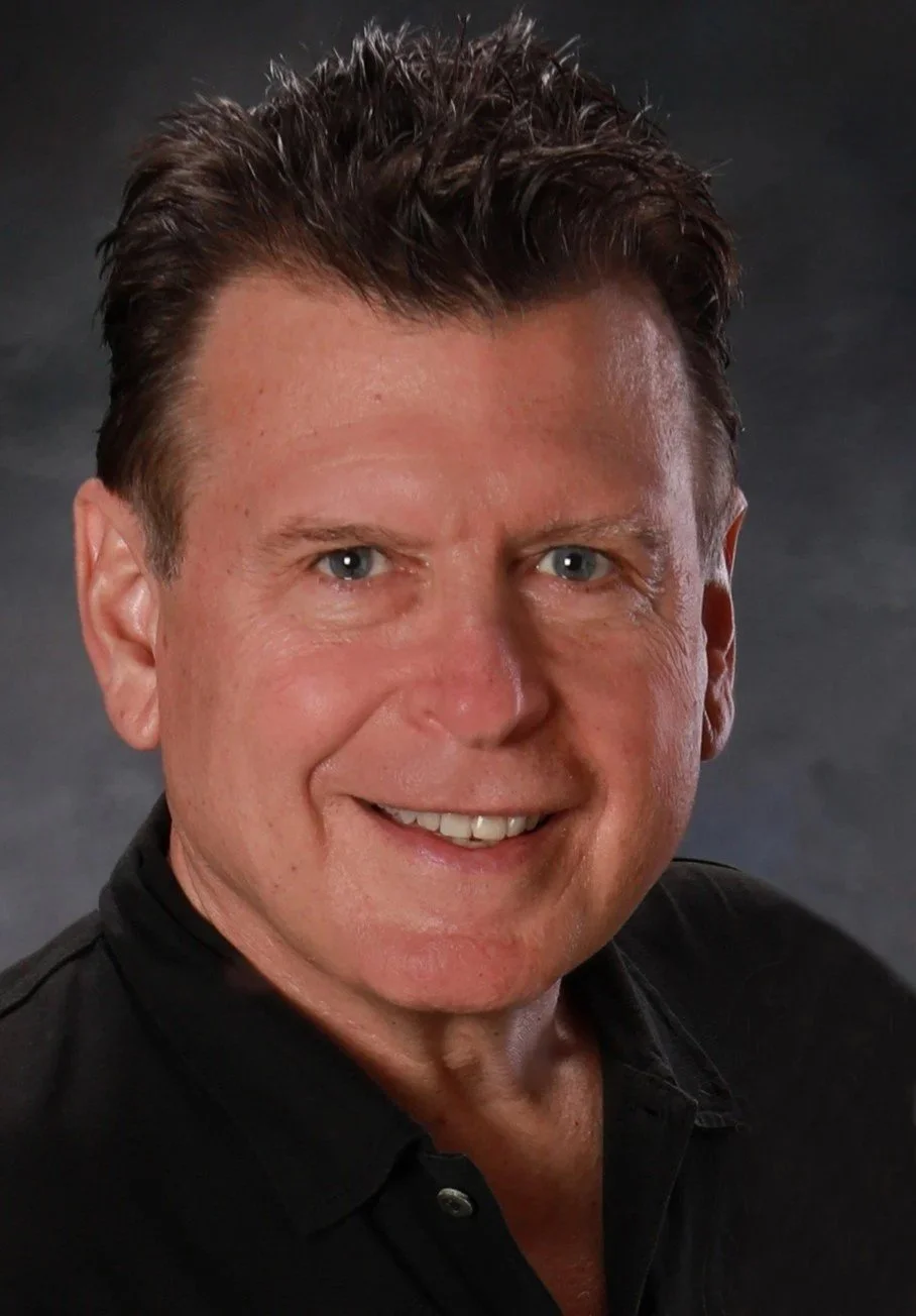 A middle-aged man with short, dark, styled hair, blue eyes, and a light complexion, smiling and wearing a black shirt, against a dark gray background.