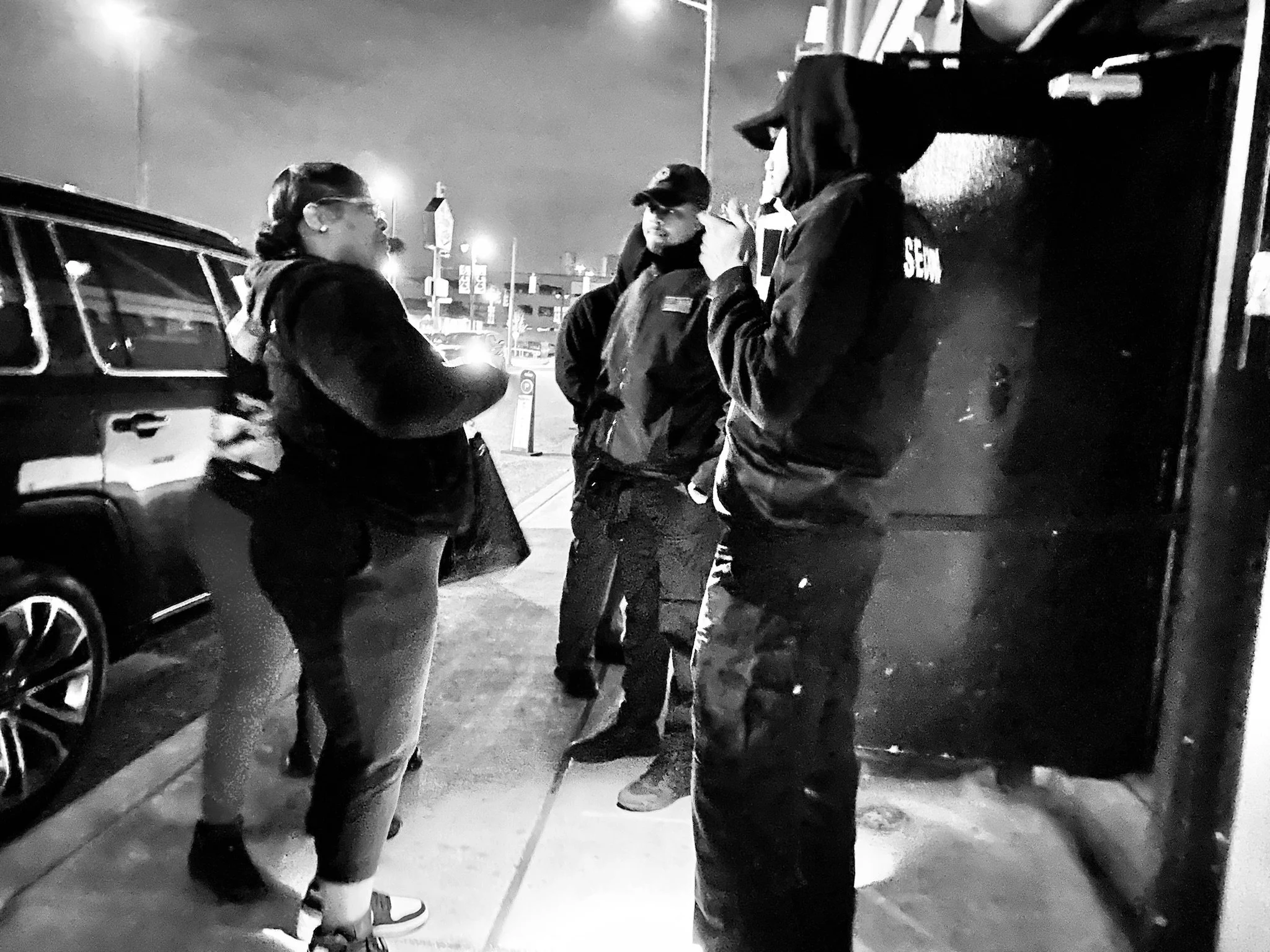 Four individuals engaging in conversation on a city sidewalk at night, with cars and streetlights in the background.