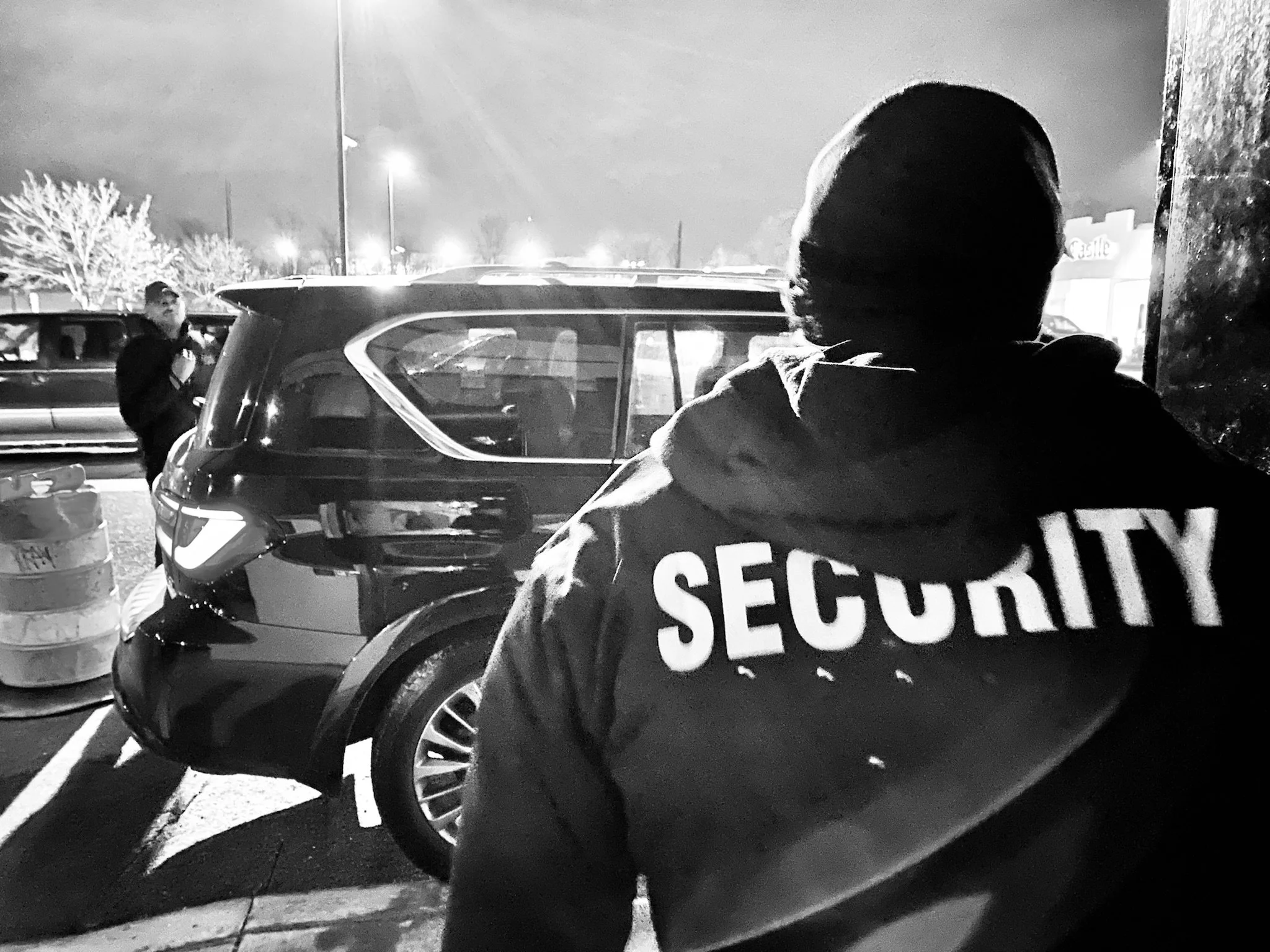 A security guard wearing a hoodie with the word 'SECURITY' on the back, standing outside at night near a parked SUV with another person in the background.
