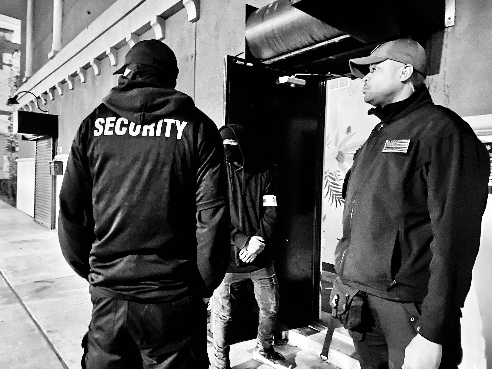 Two security guards standing outside a building, talking to a person near the entrance, at night.