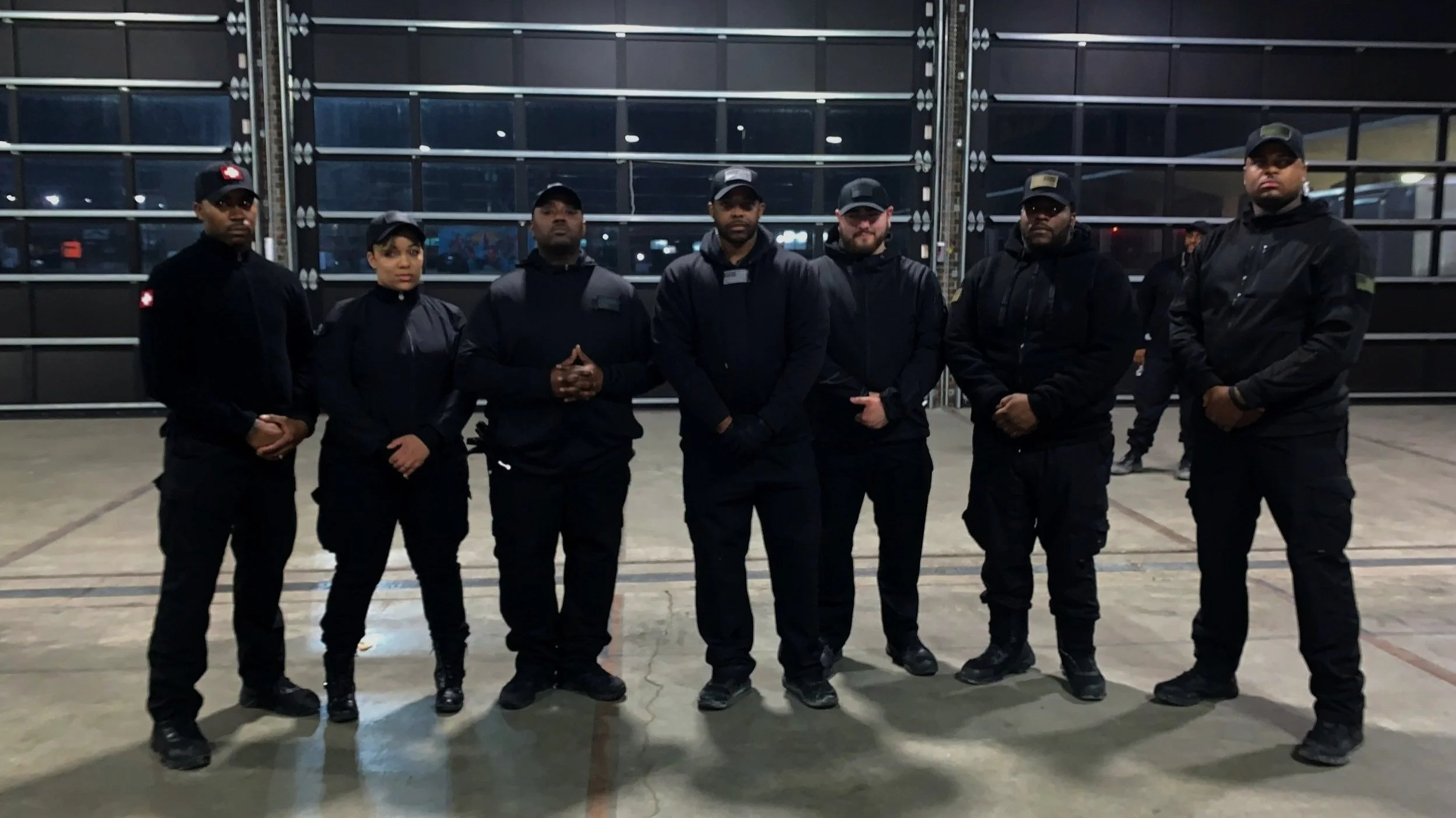 Group of seven security personnel standing in a line inside a large industrial or warehouse area at night, all dressed in black uniforms with caps.