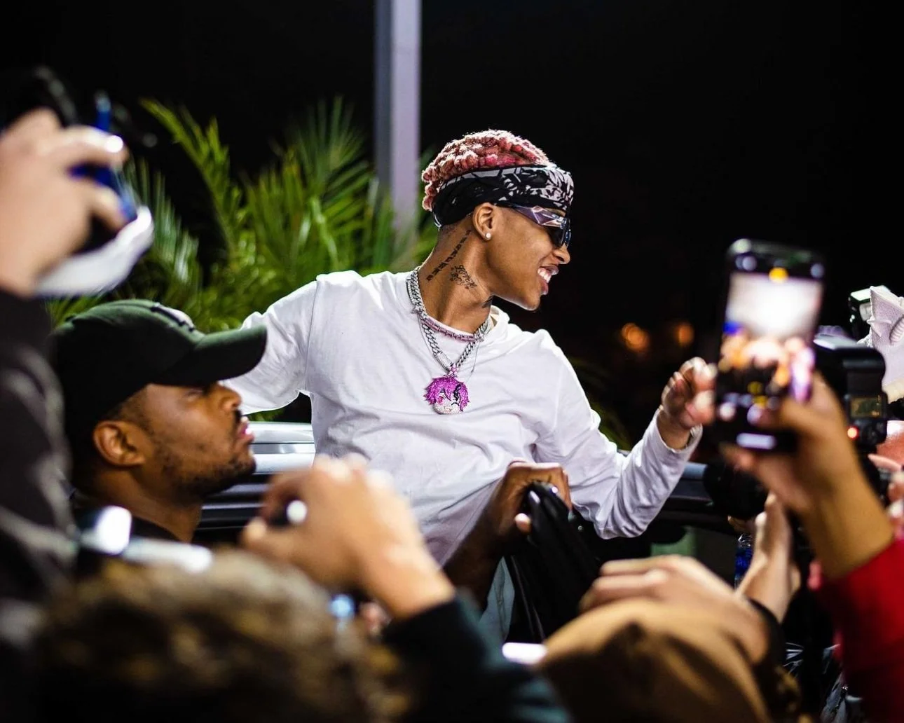 A woman with pink hair, wearing sunglasses, a headscarf, a white shirt, and jewelry, is smiling and taking a selfie with a group of people at night. There are people around her holding cameras and phones, and green plants in the background.