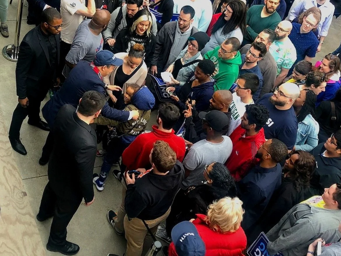 A group of people gathered in a crowded, indoor setting around a person signing autographs, with some members taking photos or videos.