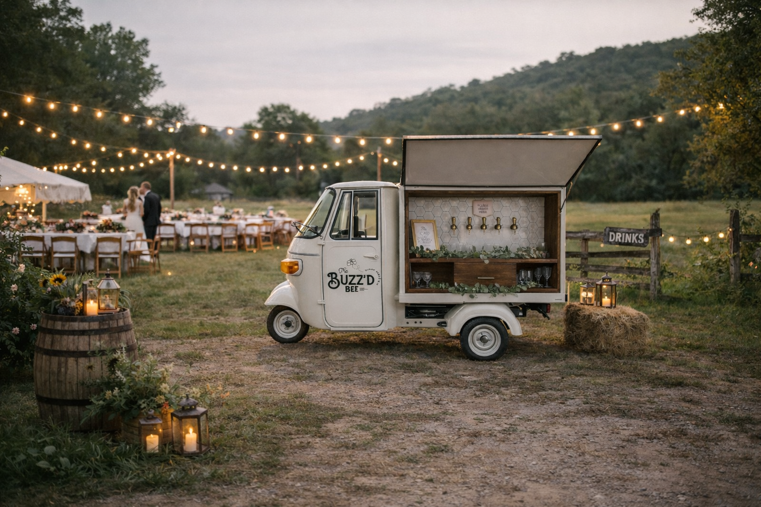 A vintage white three-wheeled food truck decorated for an outdoor event, parked on a dirt path with lanterns and hay bail decorations nearby. In the background, a tent and long banquet tables with string lights and a bride and groom are visible in a scenic outdoor setting with trees and hills.