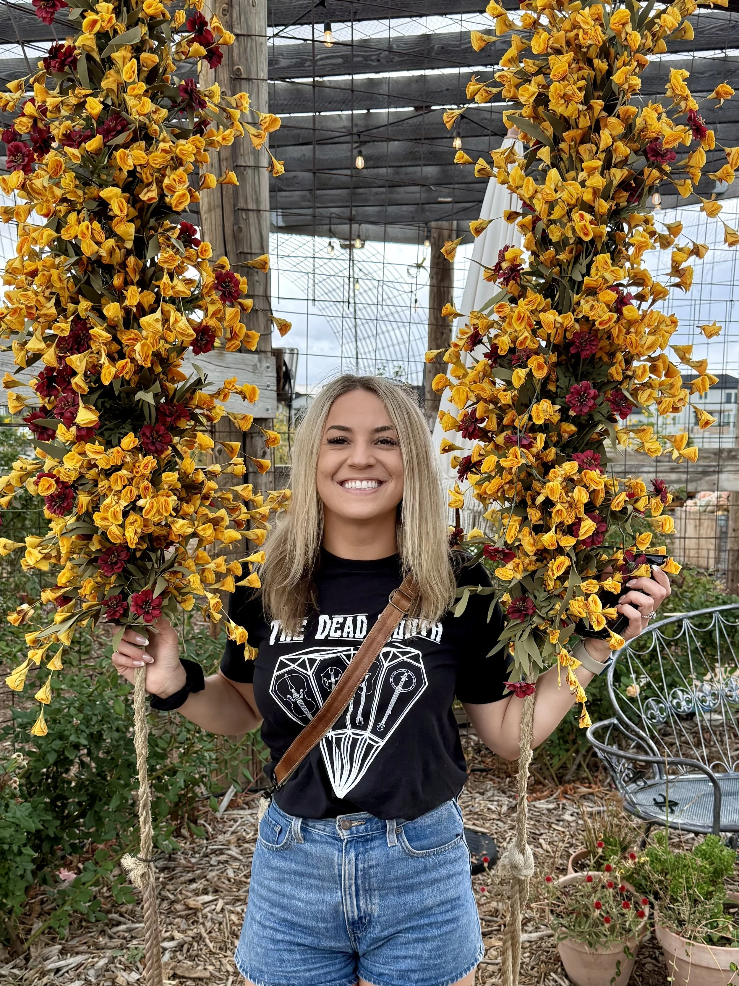 A smiling woman with blonde hair wearing a black graphic t-shirt and denim shorts, holding onto a large floral swing made of yellow and red flowers in a garden or greenhouse setting.