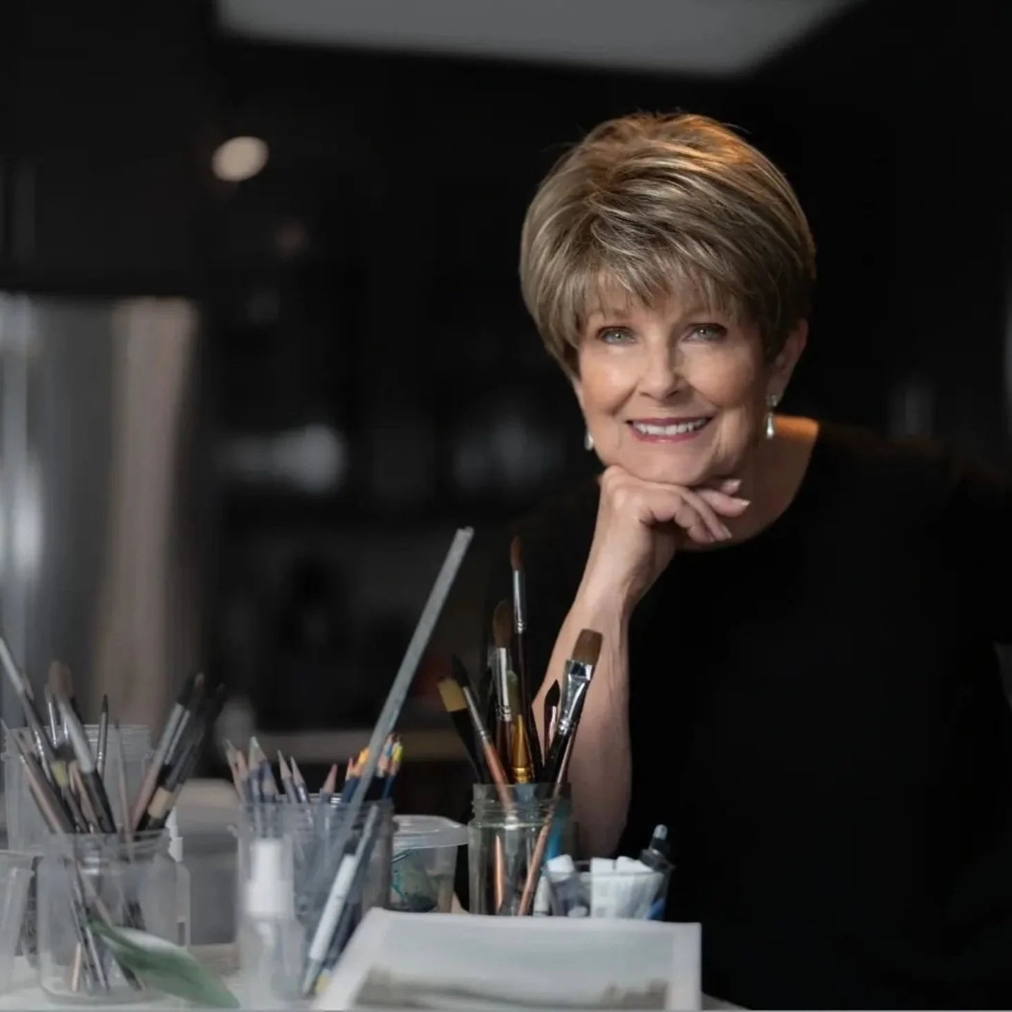 Smiling person in a dark room with chin resting on hand, surrounded by jars of paintbrushes and art supplies.
