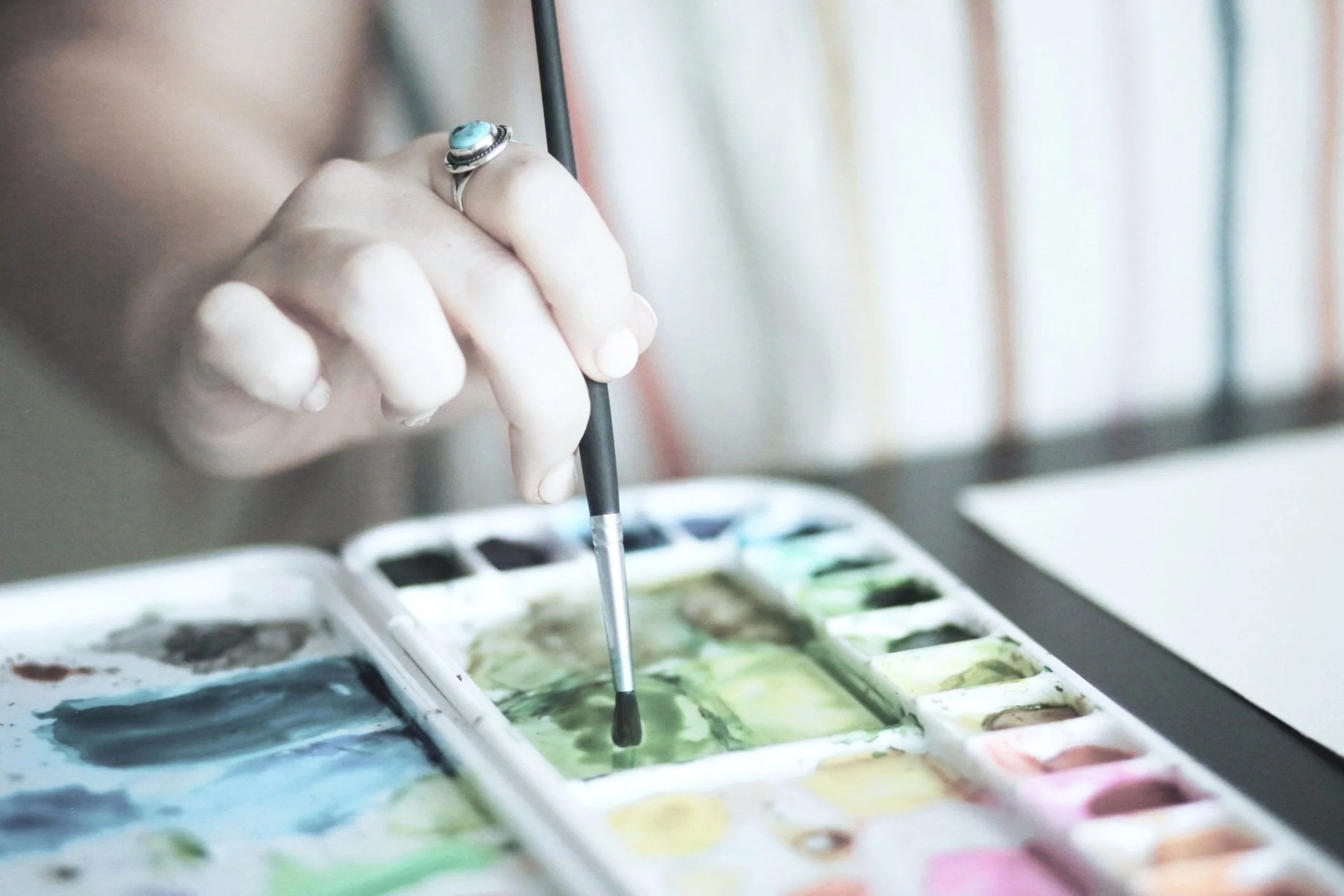 A hand holding a paintbrush over a watercolor paint palette with various colors.