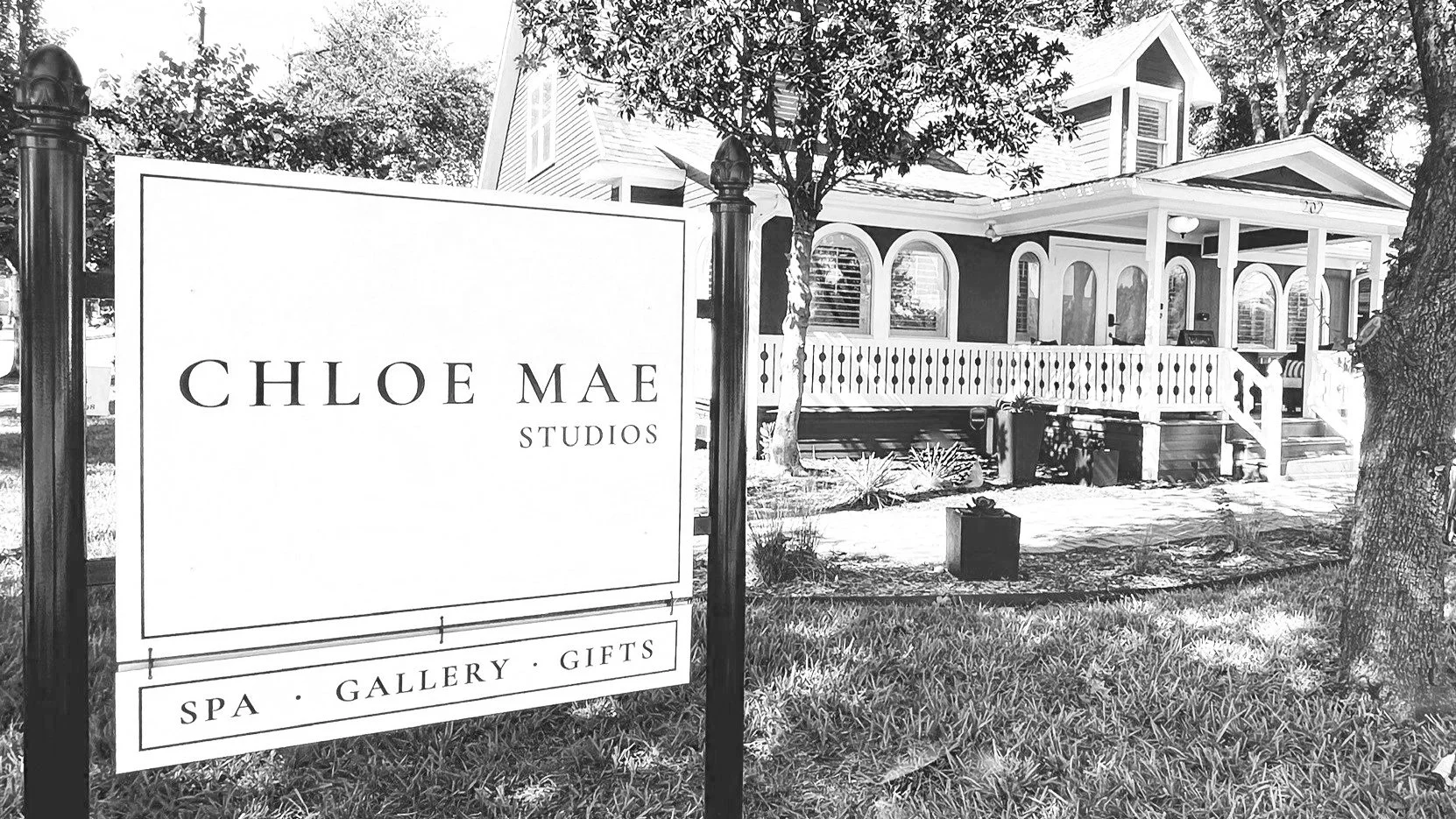 Sign reading "Chloe Mae Studios - Spa, Gallery, Gifts" next to a traditional house with a porch and trees.