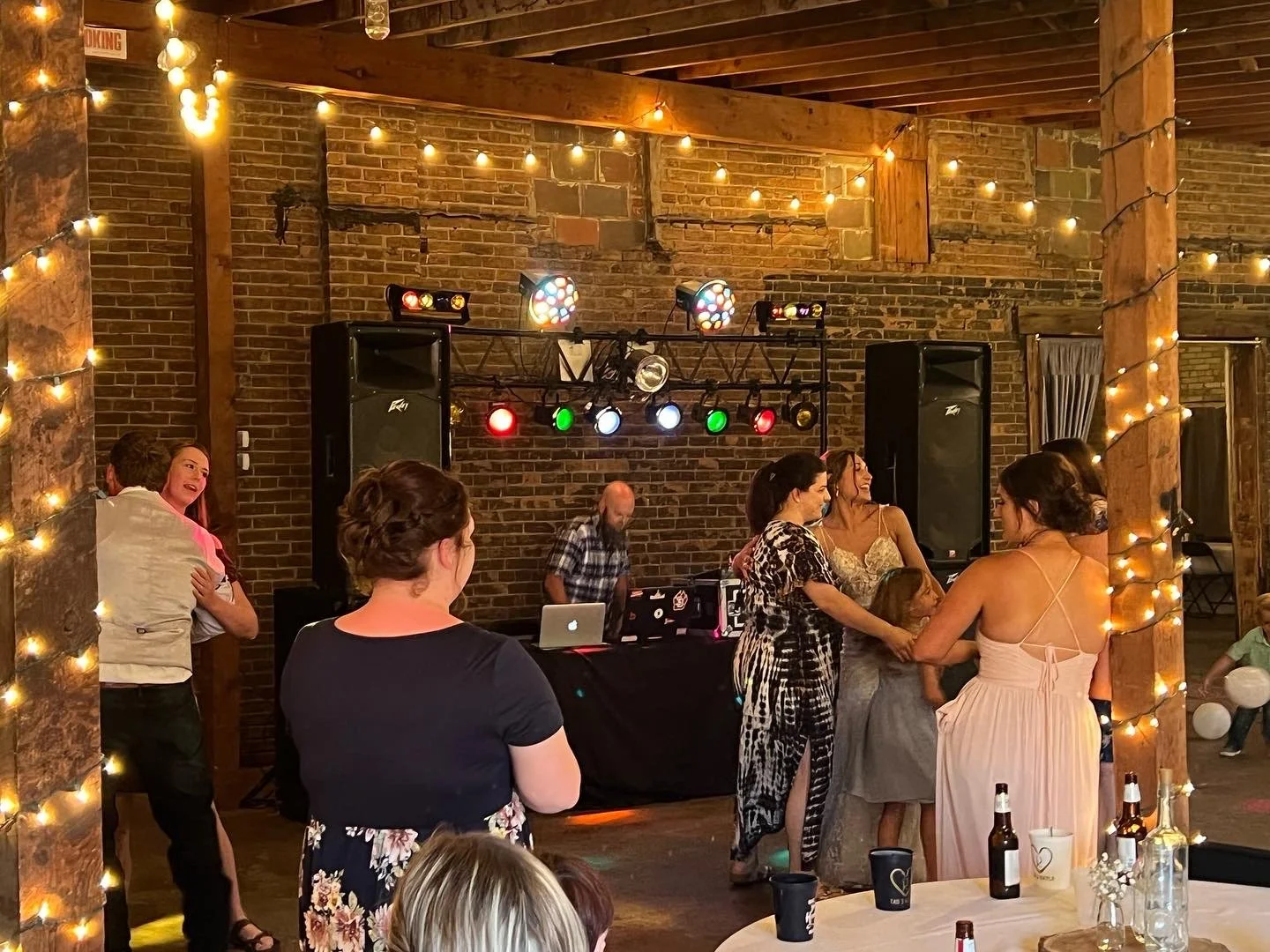 People dancing and socializing at a wedding reception with a DJ booth, disco lights, and fairy lights on brick and wood decor.