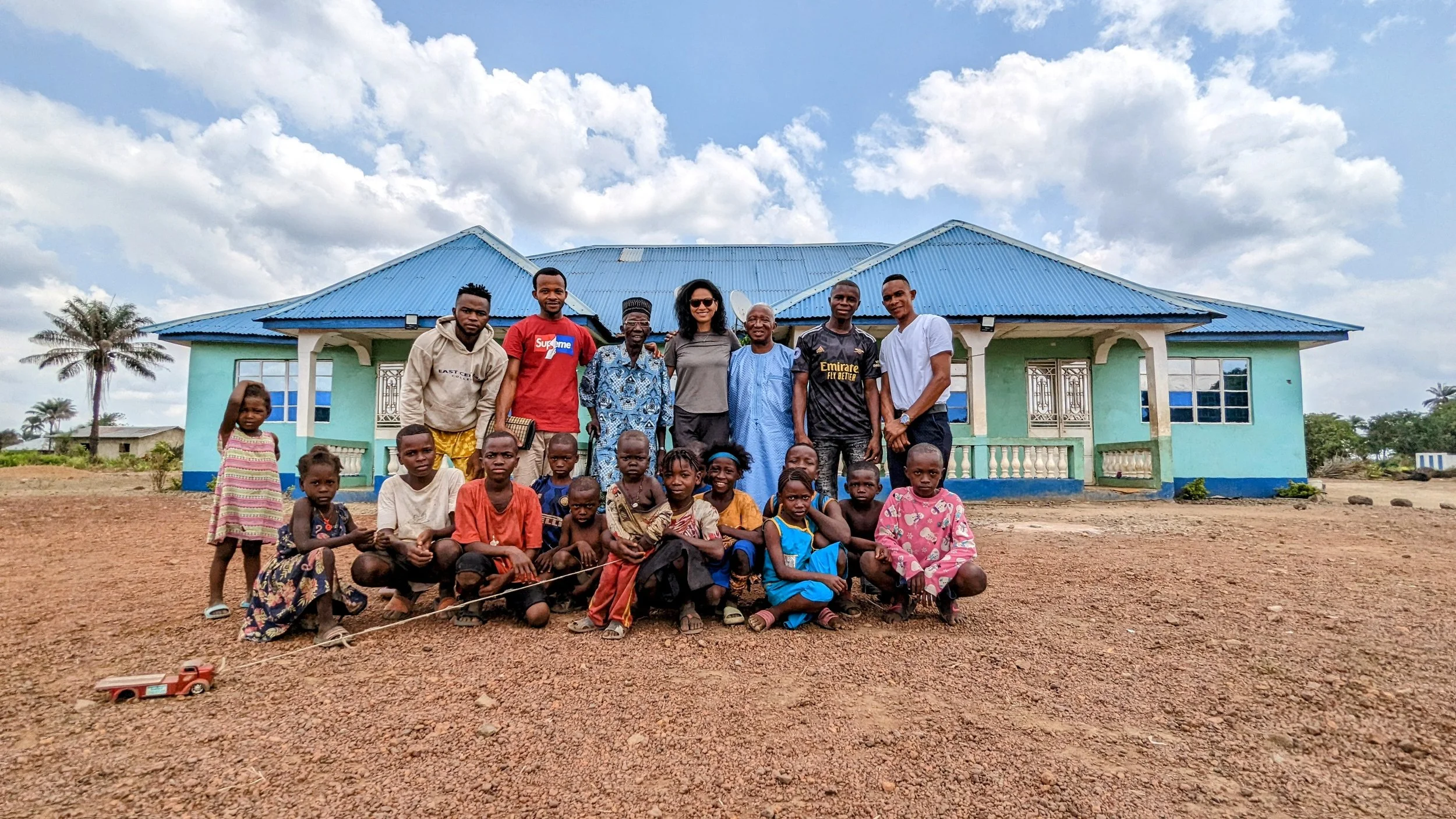 Group of children and adults standing and sitting outside a blue house with a blue roof on a dirt ground, with trees and clouds in the background.
