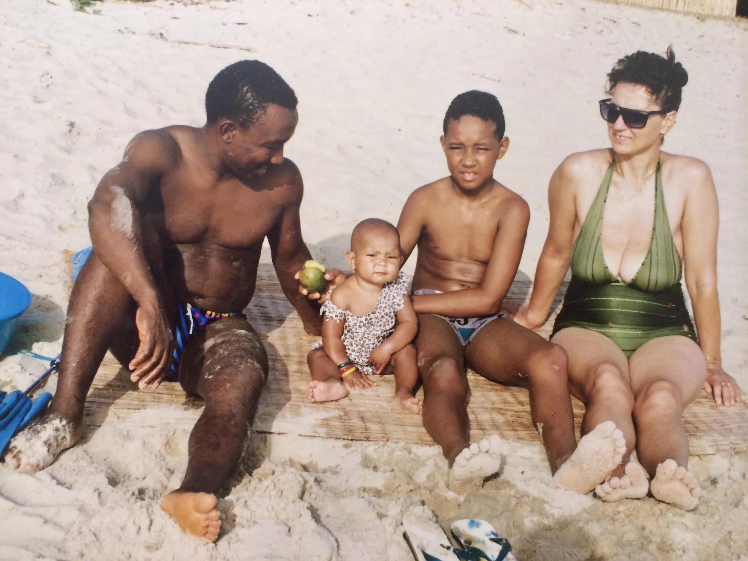 Four people sitting on a beach: a man, a woman, and two children. The man and boy are shirtless, and the woman is wearing sunglasses and a green swimsuit. They are sitting on a wooden mat with sand around them, and the man is holding an apple.