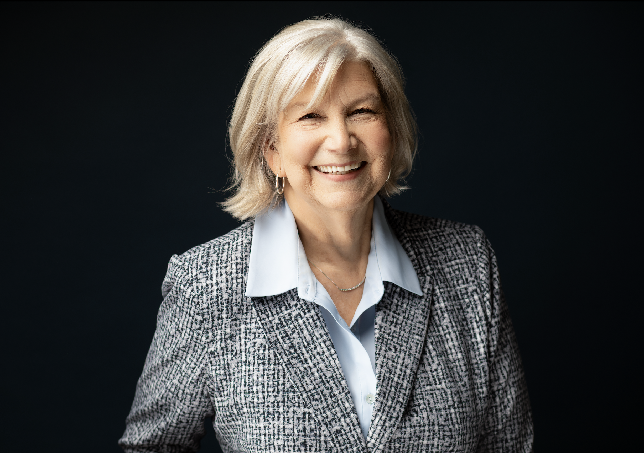 A smiling elderly woman with short blonde hair, wearing a light blue collared shirt and a patterned blazer, standing against a dark background.