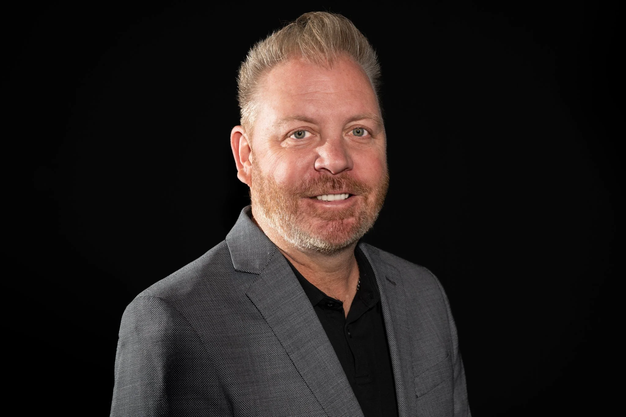 Portrait of a man with light hair and beard, smiling, wearing a gray blazer over a black shirt, against a black background.