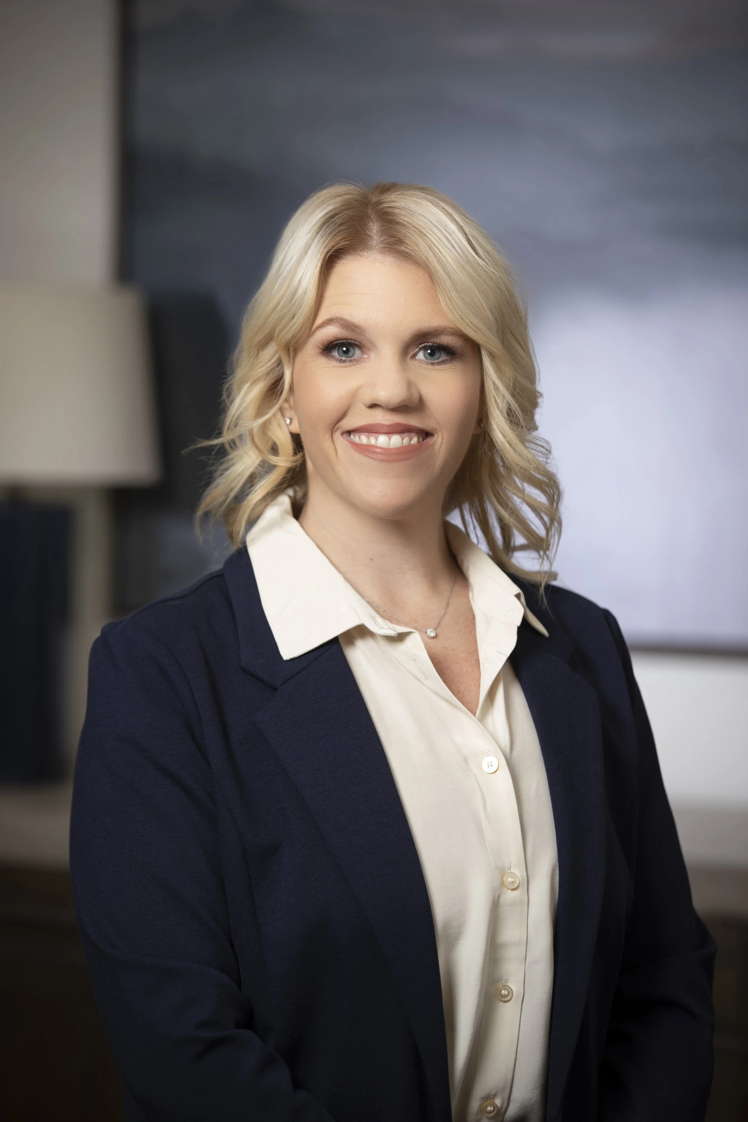 A professional woman with blonde, shoulder-length hair, wearing a cream blouse and navy blazer, smiling in a modern office setting.