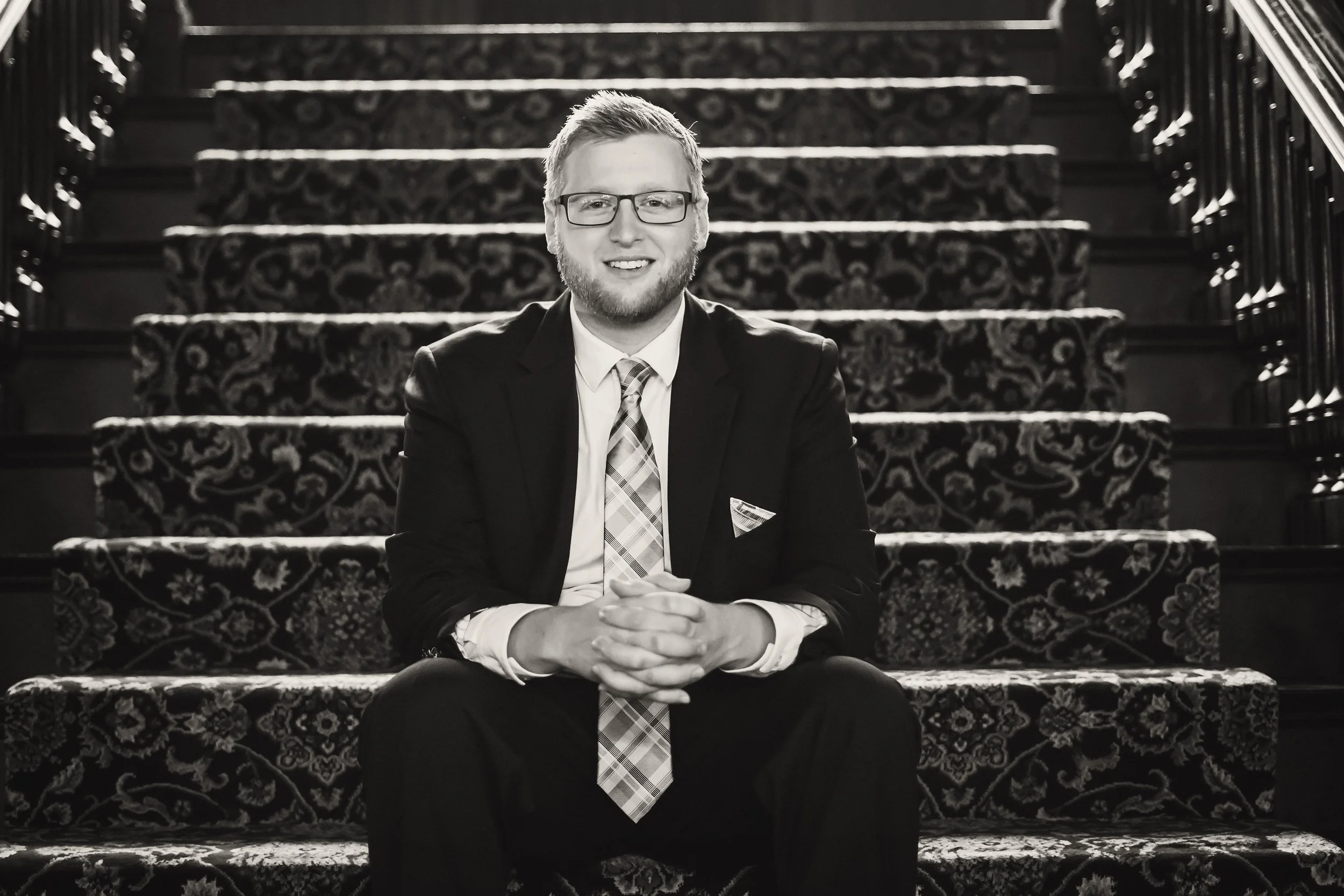 A man wearing glasses, a suit, and a tie sitting on a staircase with a patterned carpet, smiling at the camera.