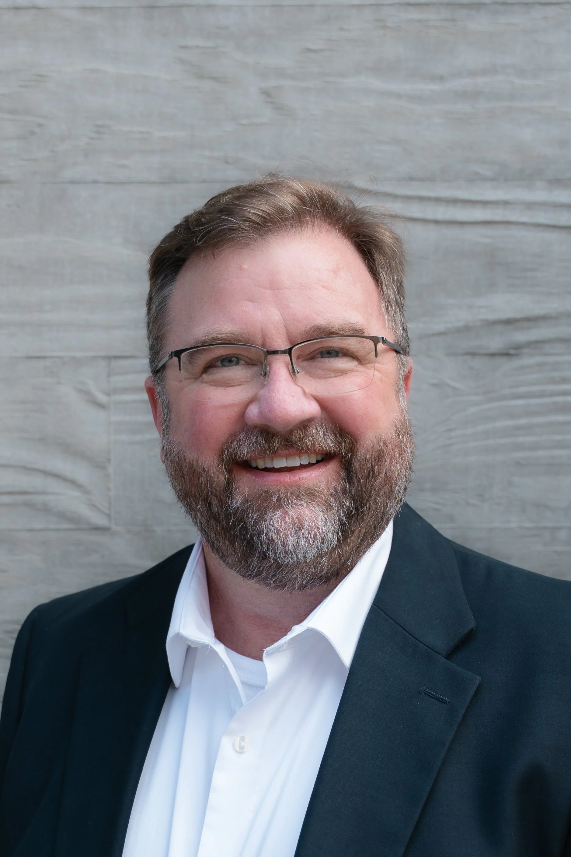 A smiling man with glasses, a beard, and short hair, wearing a white shirt and black blazer, standing in front of a light-colored wooden wall.