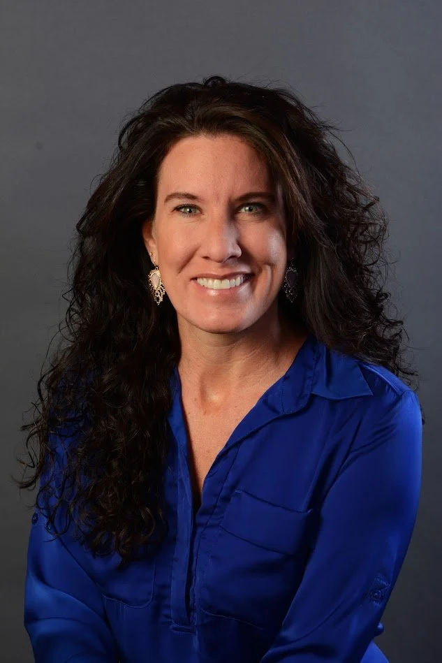 A woman with long, curly dark hair wearing a bright blue blouse and gold earrings, smiling against a gray background.